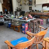 A cluttered garage sale scene: tables laden with items, chairs, and a variety of goods.