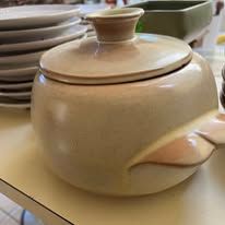 Tan ceramic lidded serving bowl with a rounded shape.  A stack of plates sits in the background.