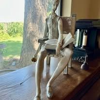 Statue of a slender figure sitting, reading a book indoors, near a window.