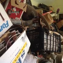 Cluttered room: boxes, bags, baskets, and hats. Overstuffed with belongings, in a dimly lit setting.