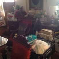 Cluttered room with furniture, boxes, and various items, suggesting a storage area or a room in disarray.