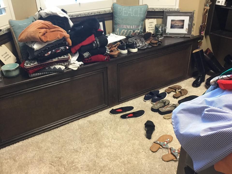 Pile of clothes and shoes on a bench and floor in a room with a beige carpet.