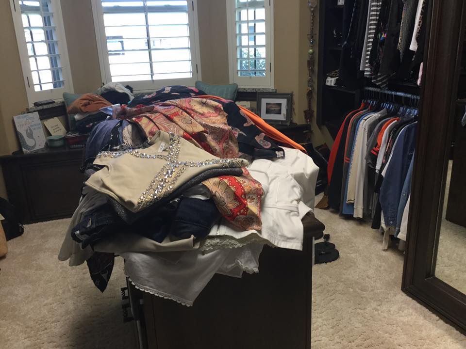 Clothes piled on a chest in a bedroom. Closets with hanging clothes, large mirror, windows in background.