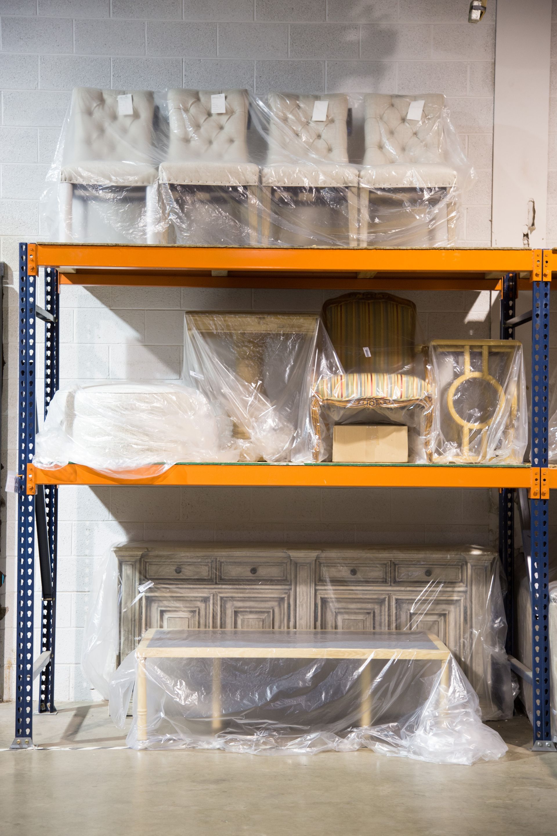 Shelving unit with furniture covered in plastic wrap; chairs, tables, and a buffet in a warehouse.