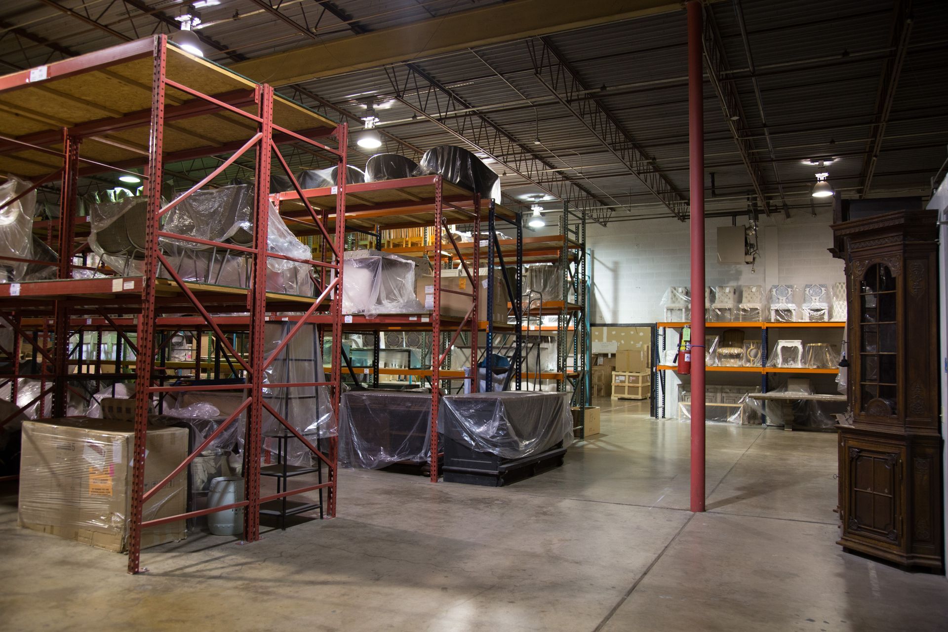 Warehouse interior with red shelving units, packed boxes, and furniture, with a red support pole in the middle.