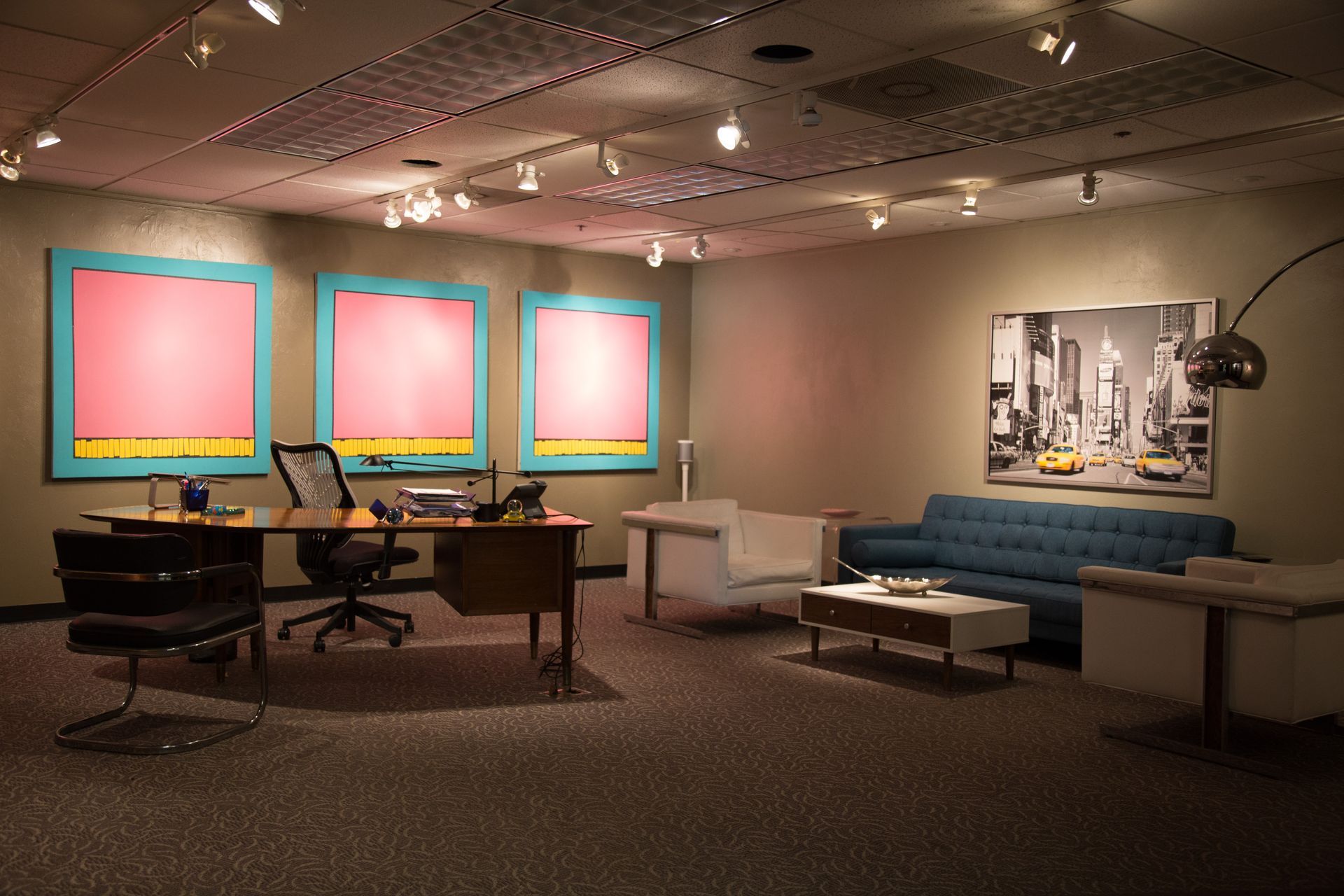 Office with desk, seating area, artwork; pink and blue accents, neutral walls and carpet.