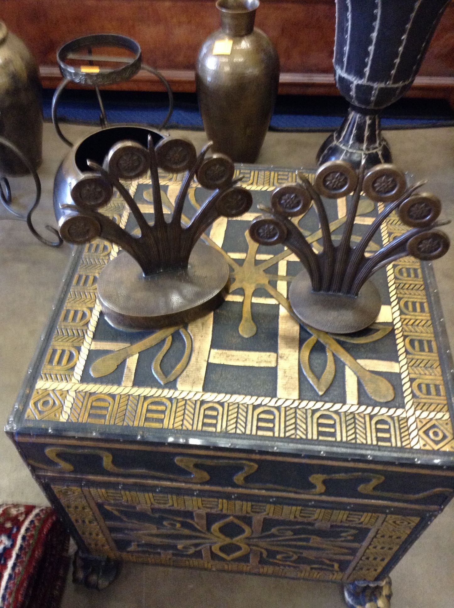 Two bronze decorative pieces on a black and gold patterned chest, with vases in the background.