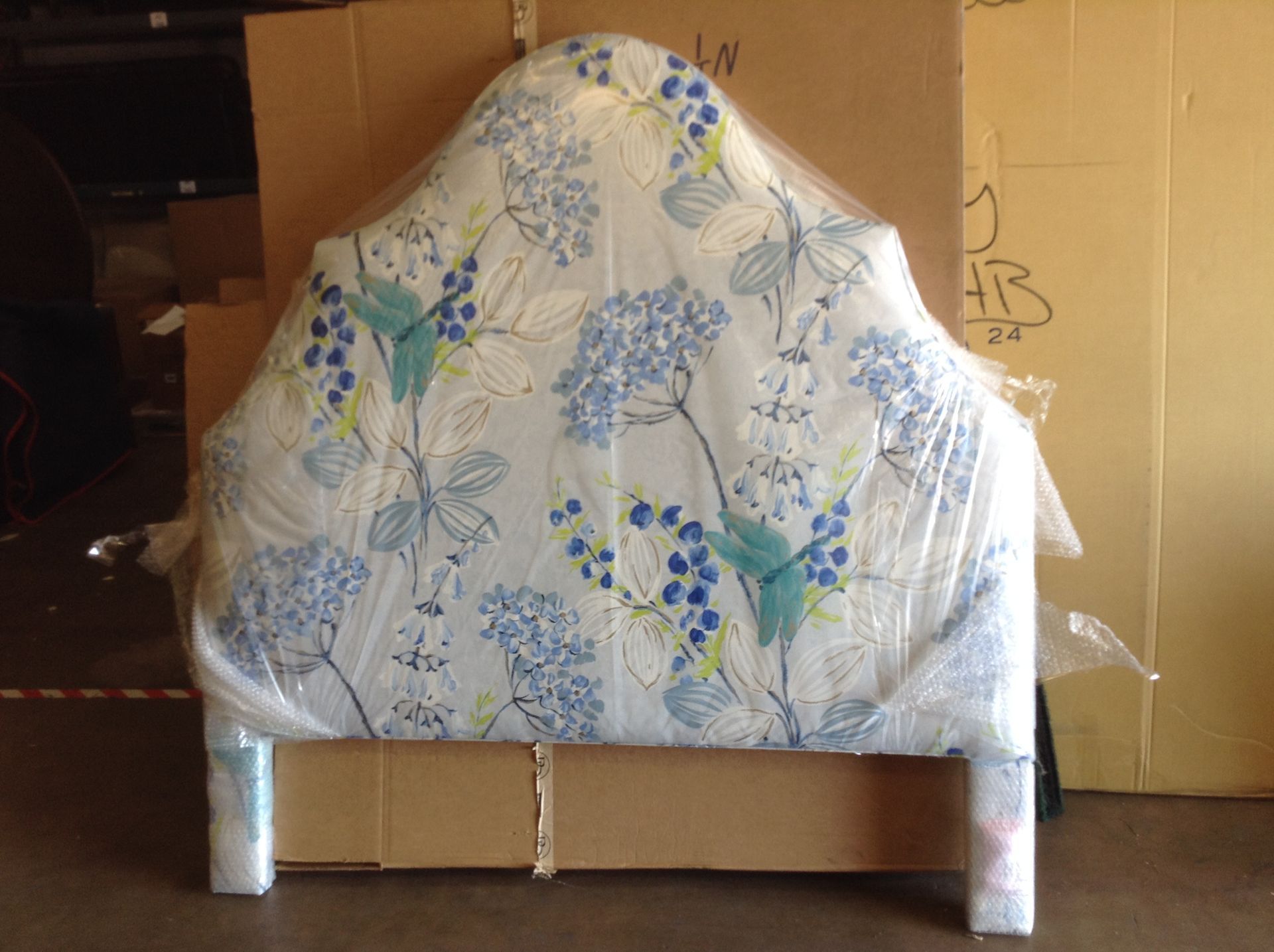 Upholstered headboard with a floral pattern, wrapped in plastic, in front of cardboard boxes.