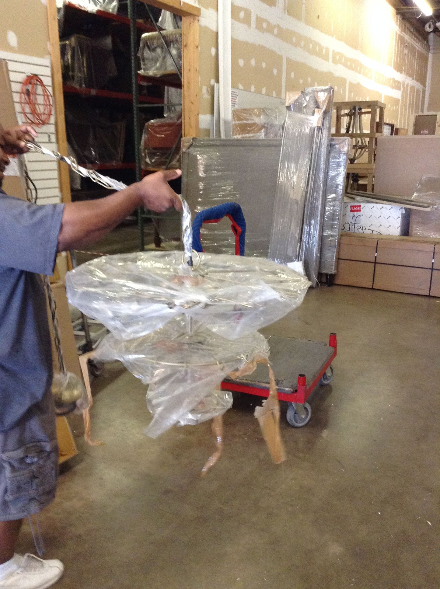 Man holding wrapped furniture in warehouse, standing by a cart.
