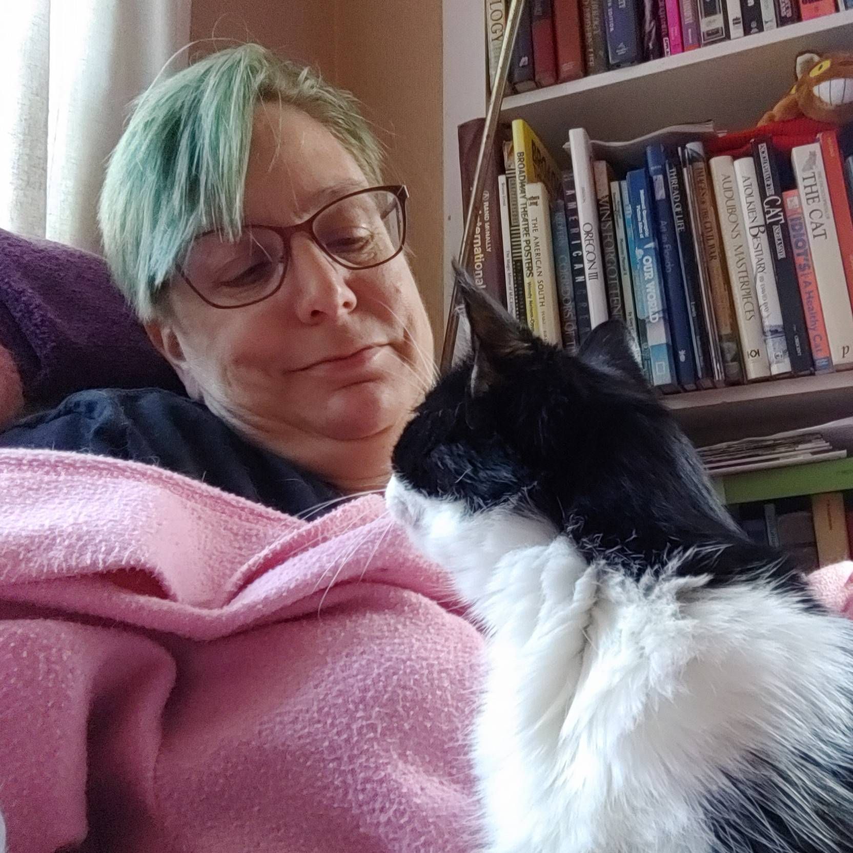 A woman with green hair is holding a black and white cat on her lap.