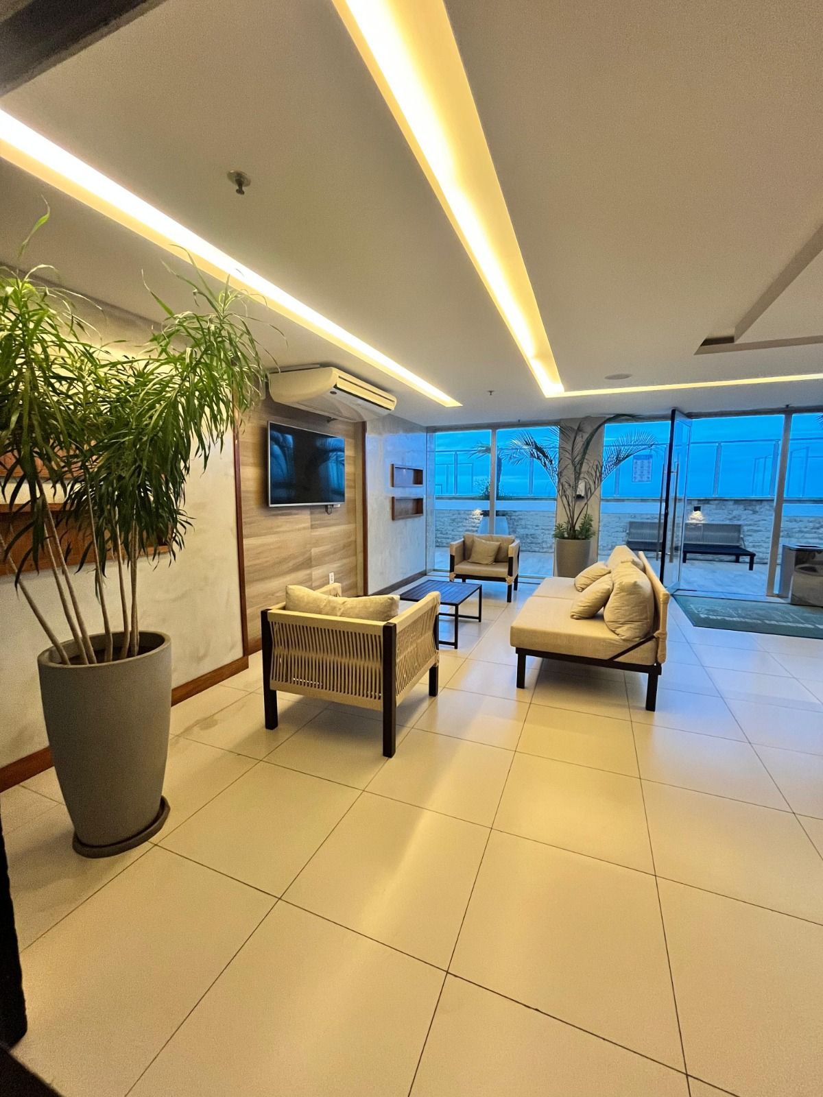 Salão de recepção moderno com sofás, planta em vaso, TV e janelas panorâmicas com vista para a cidade.