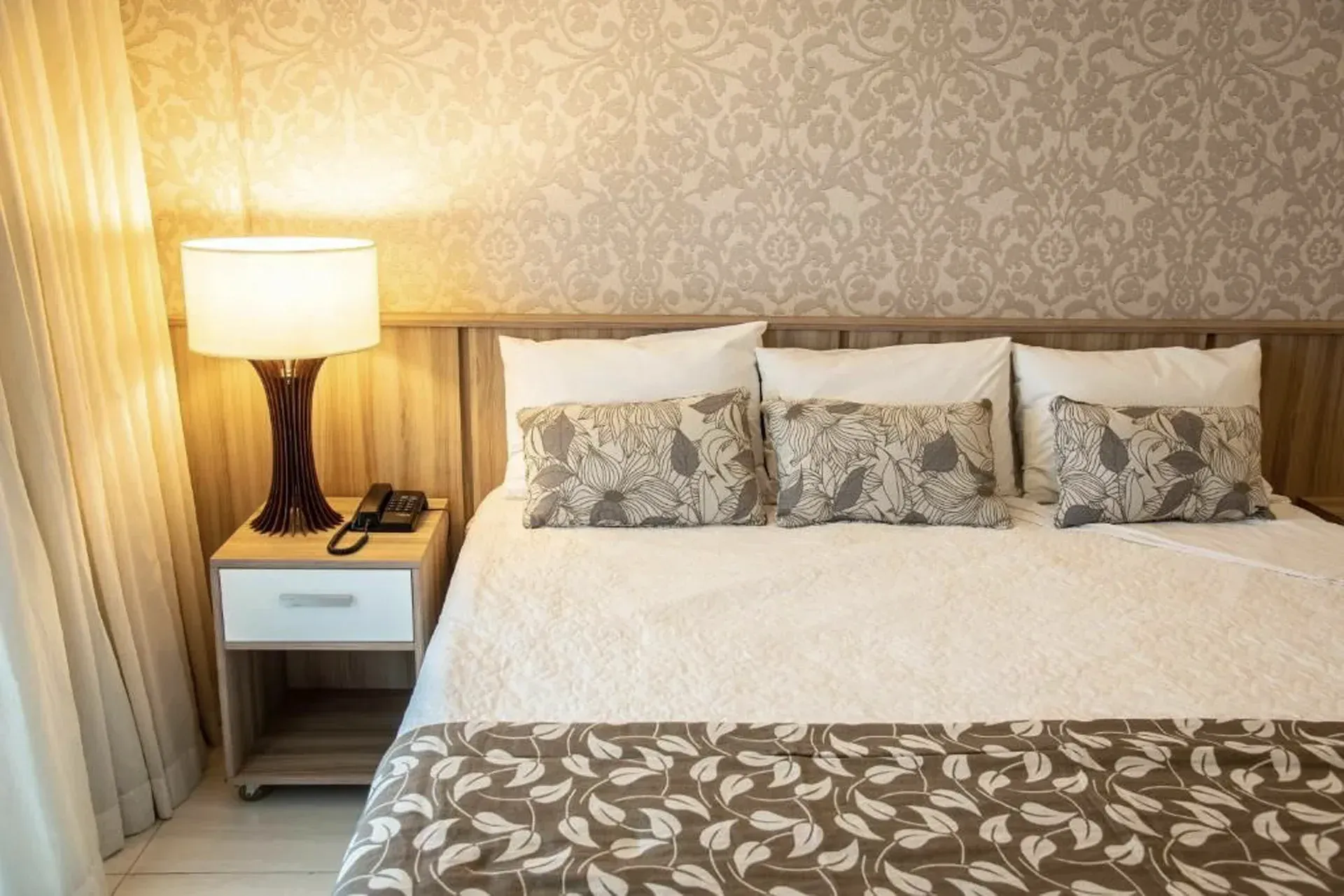 Quarto com cama arrumada, abajur e papel de parede estampado em iluminação aconchegante.