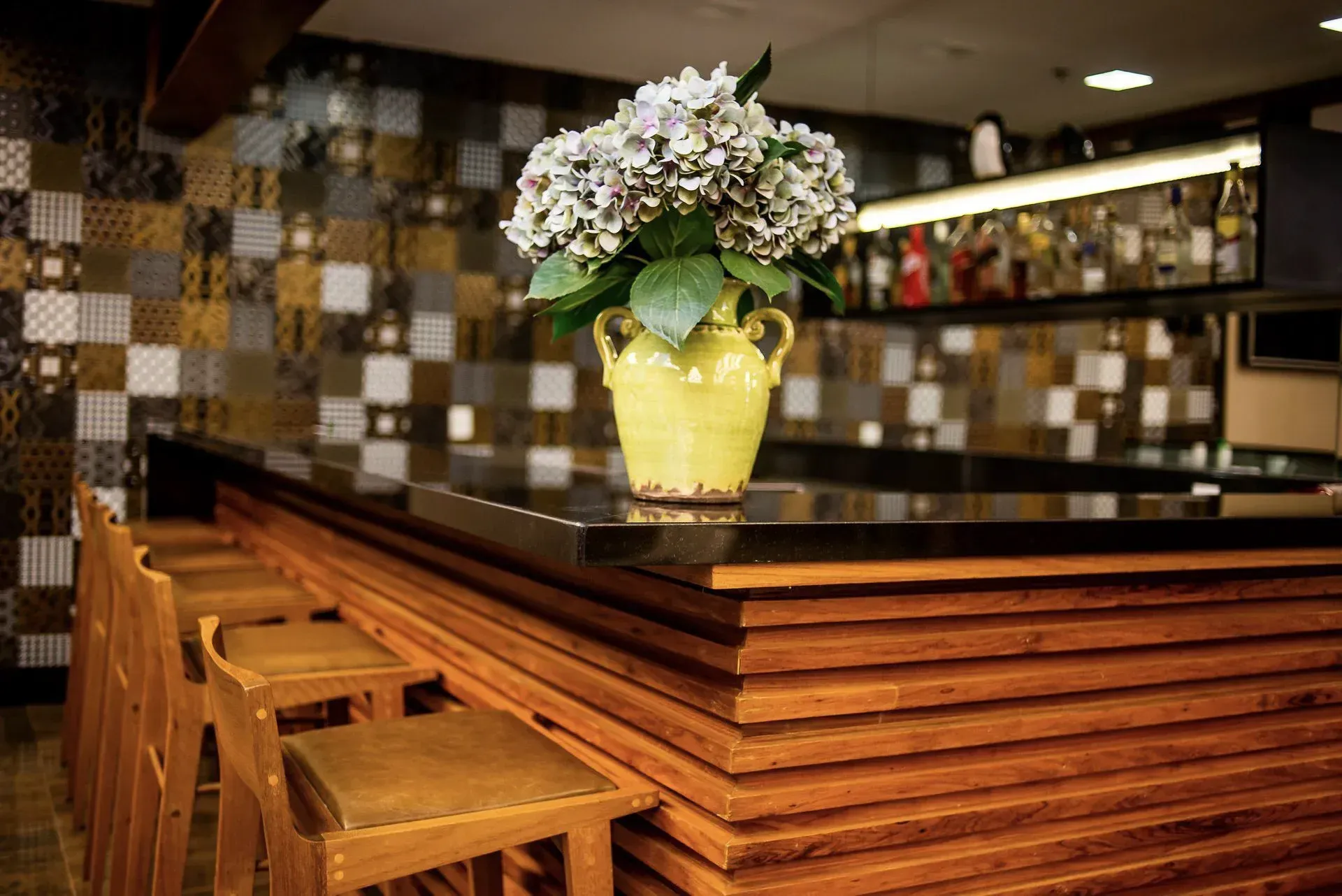 Balcão de bar de madeira com banquetas e um vaso de flores amarelo em um interior revestido de azulejos.