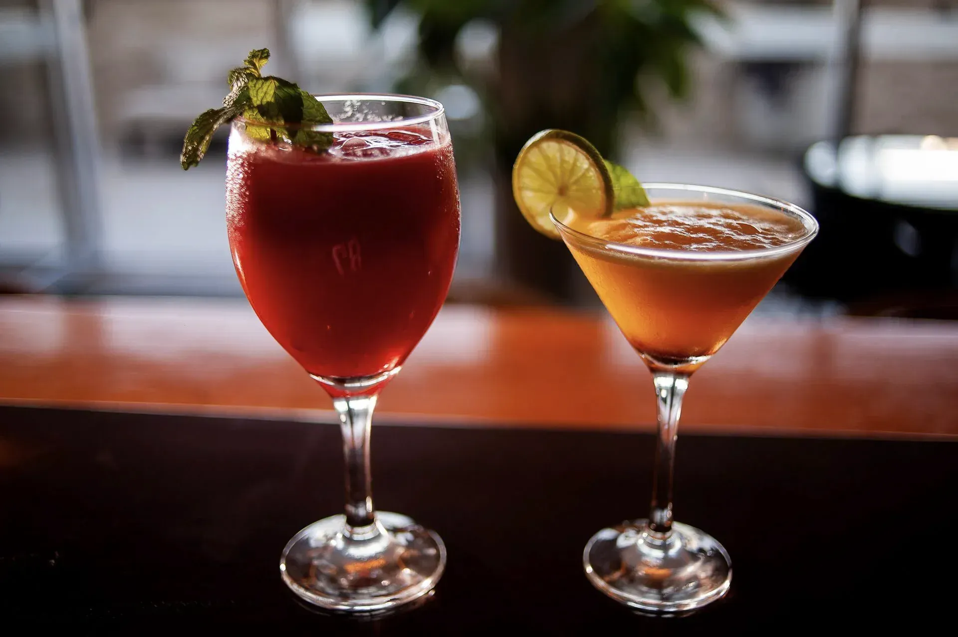Dois coquetéis coloridos em um bar, um vermelho com hortelã e um laranja com uma fatia de limão.