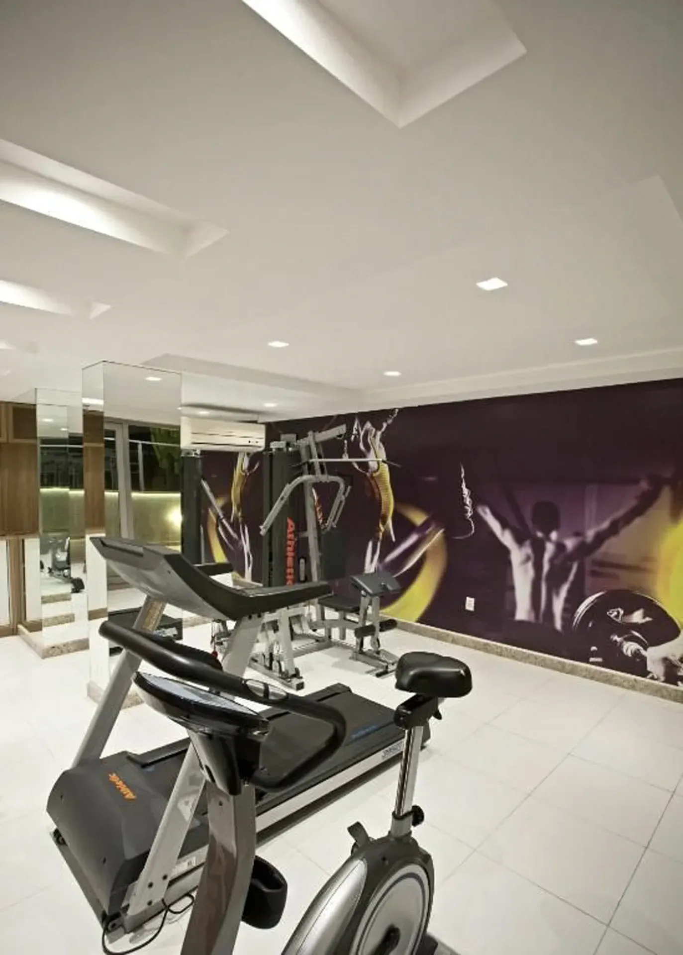 Academia interna com bicicletas ergométricas e equipamentos de musculação em frente a uma parede com um mural roxo.