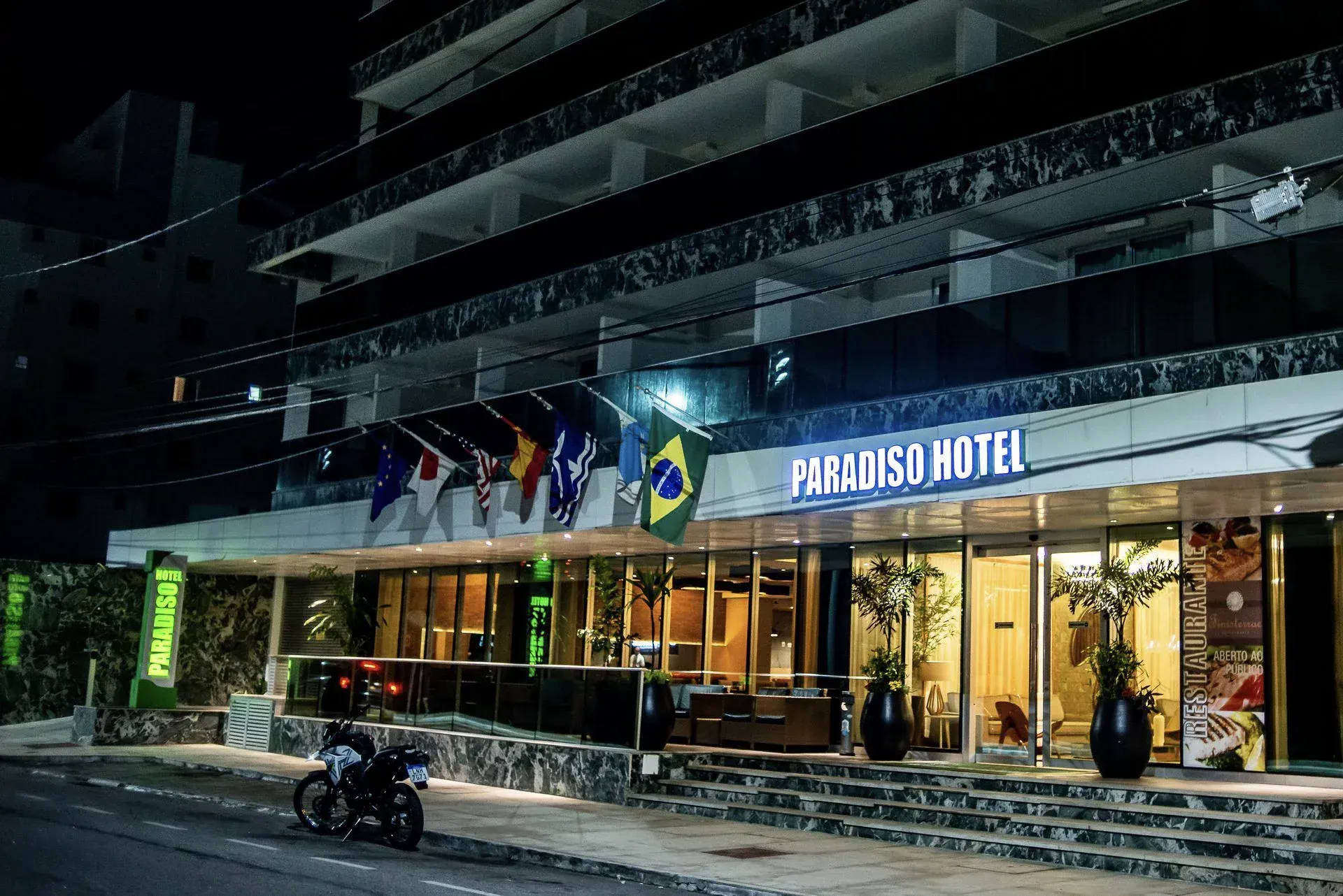 Vista noturna da entrada do Hotel Paros com toldo iluminado, portas de vidro e motocicleta estacionada.