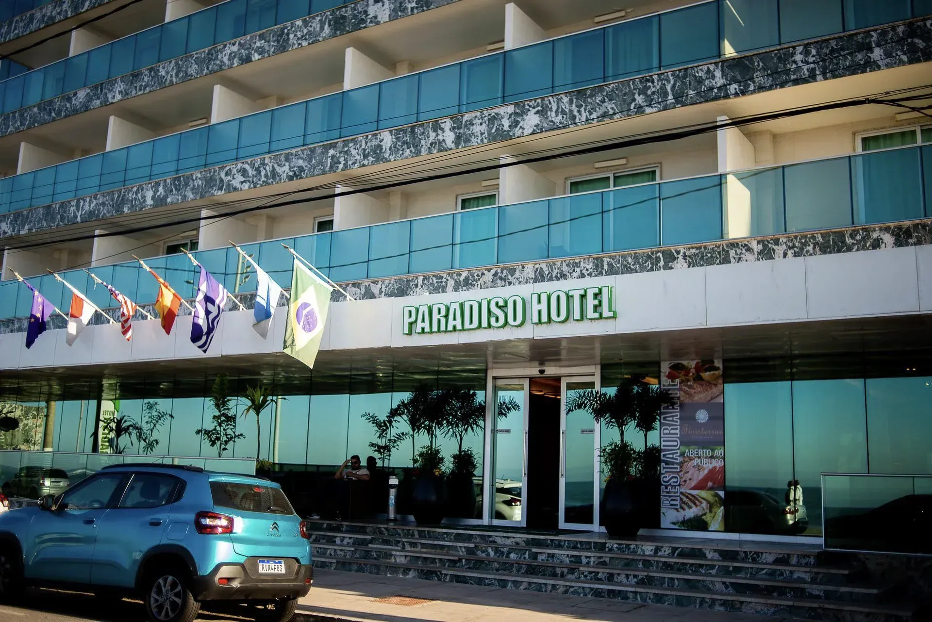 Exterior do Hotel Paradiso com bandeiras, fachada de vidro e carro azul estacionado na entrada.