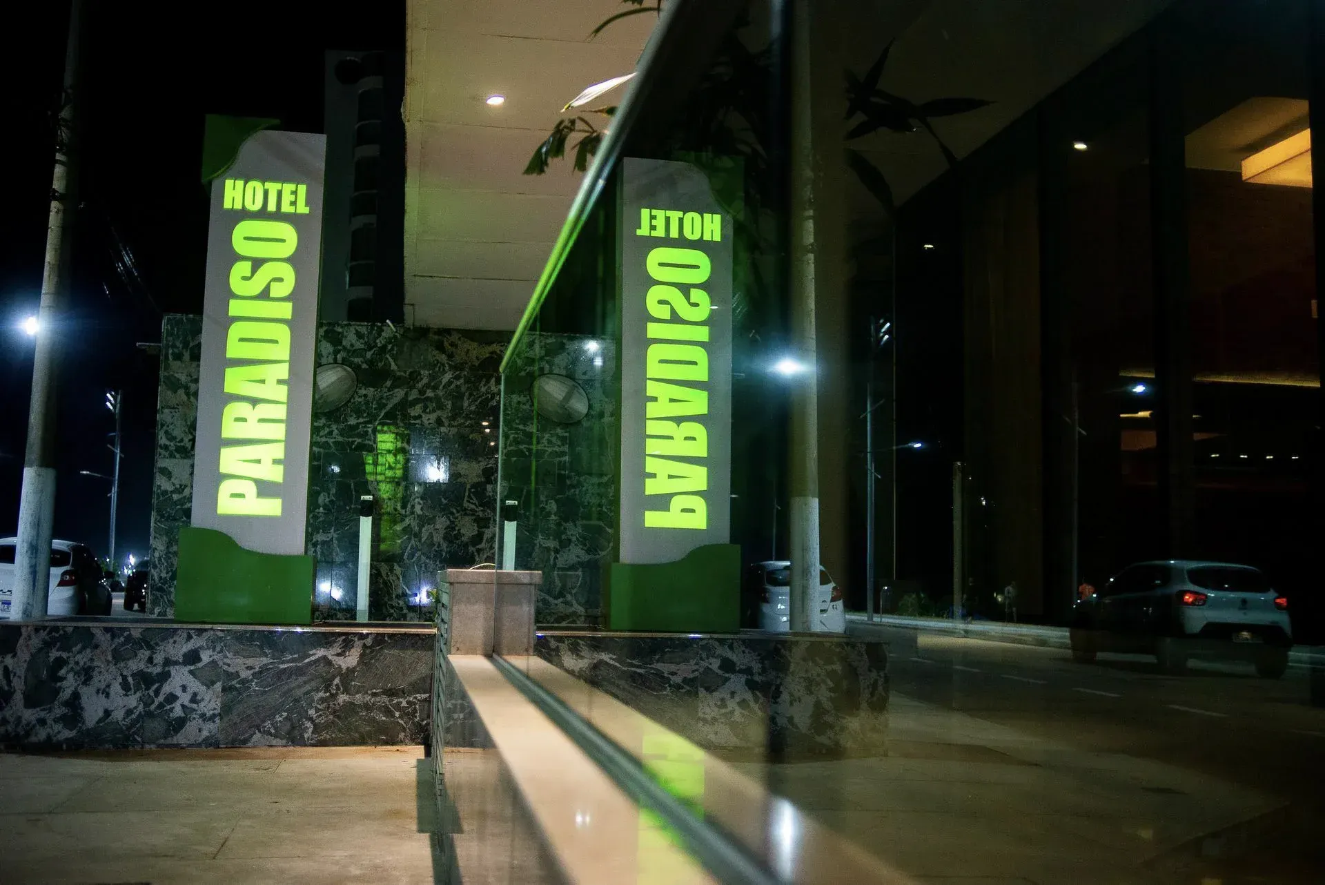 Entrada noturna do hotel com pilares iluminados em verde com os dizeres “Paradiso” e “L&BVARDO” ao lado de portas de vidro.