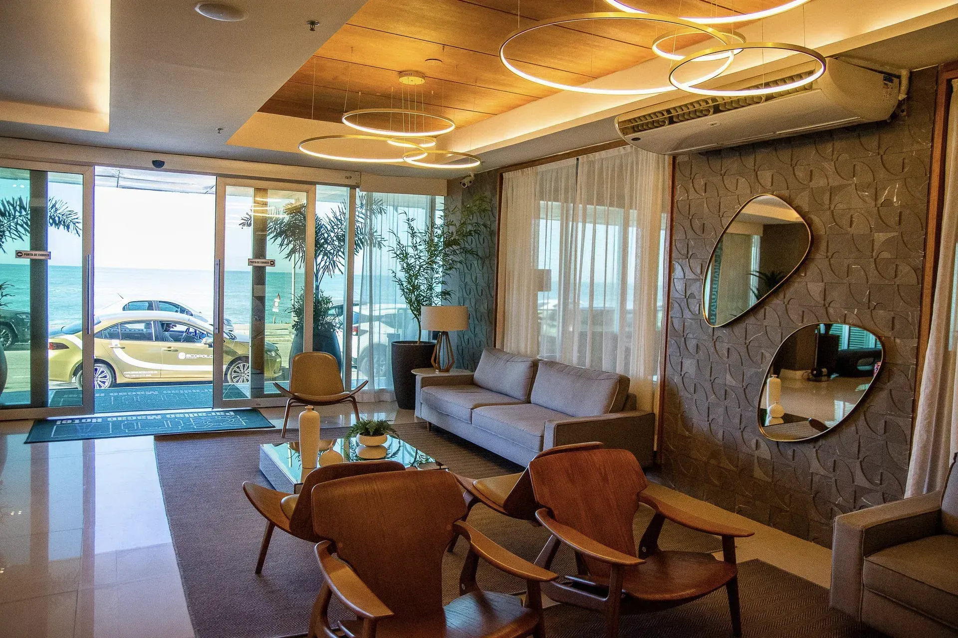 Sala de estar moderna com sofá, cadeiras de madeira, parede espelhada e grandes janelas com vista para um pátio e para o mar.