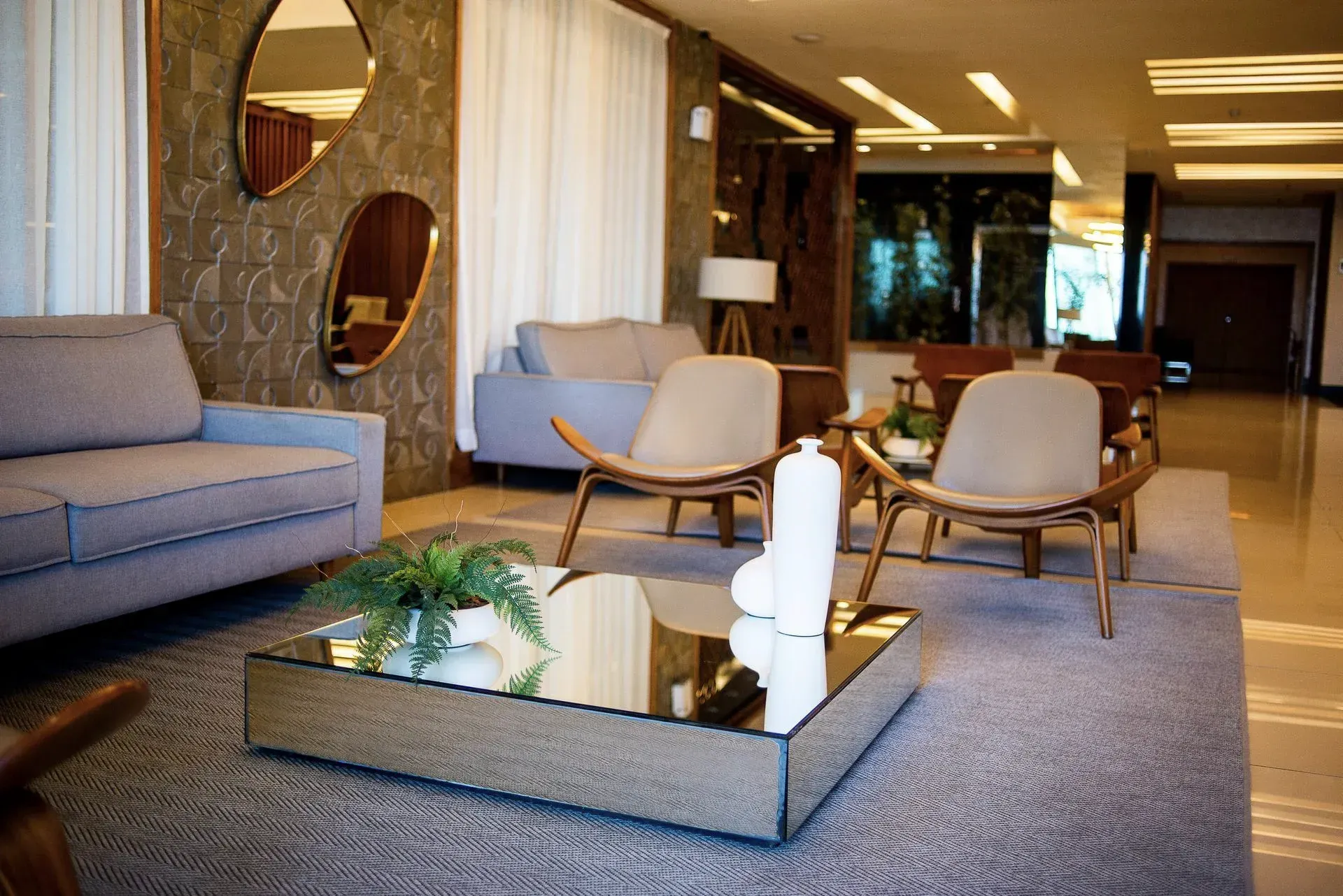 Sala de estar moderna com sofás cinza, cadeiras de madeira, mesa de centro de vidro e tapete estampado.