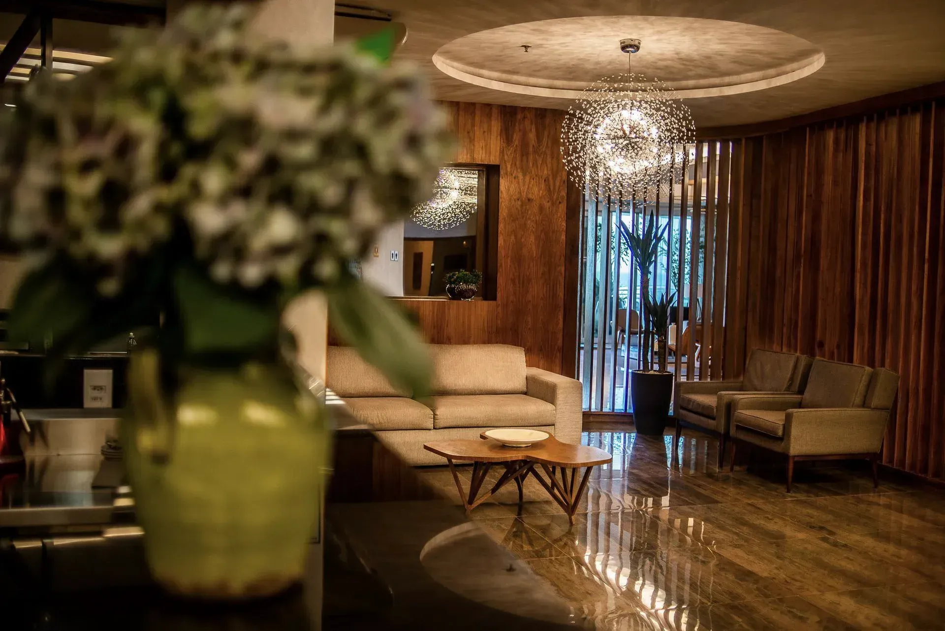 Sala de estar elegante de hotel com sofás bege, lustre, piso polido e flores desfocadas em primeiro plano.