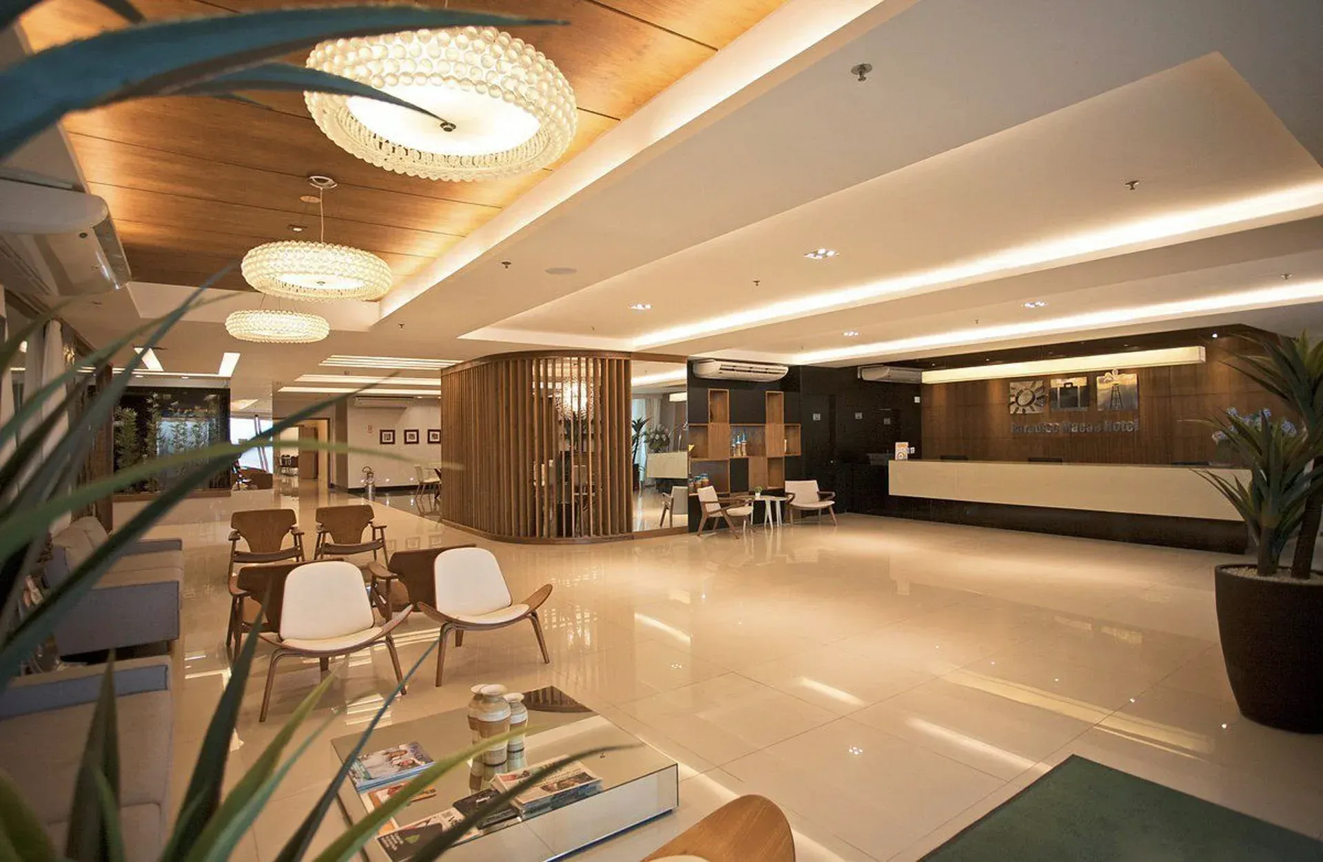 Lobby de hotel moderno com pisos polidos, cadeiras brancas, iluminação aconchegante e área de estar com painéis de madeira.