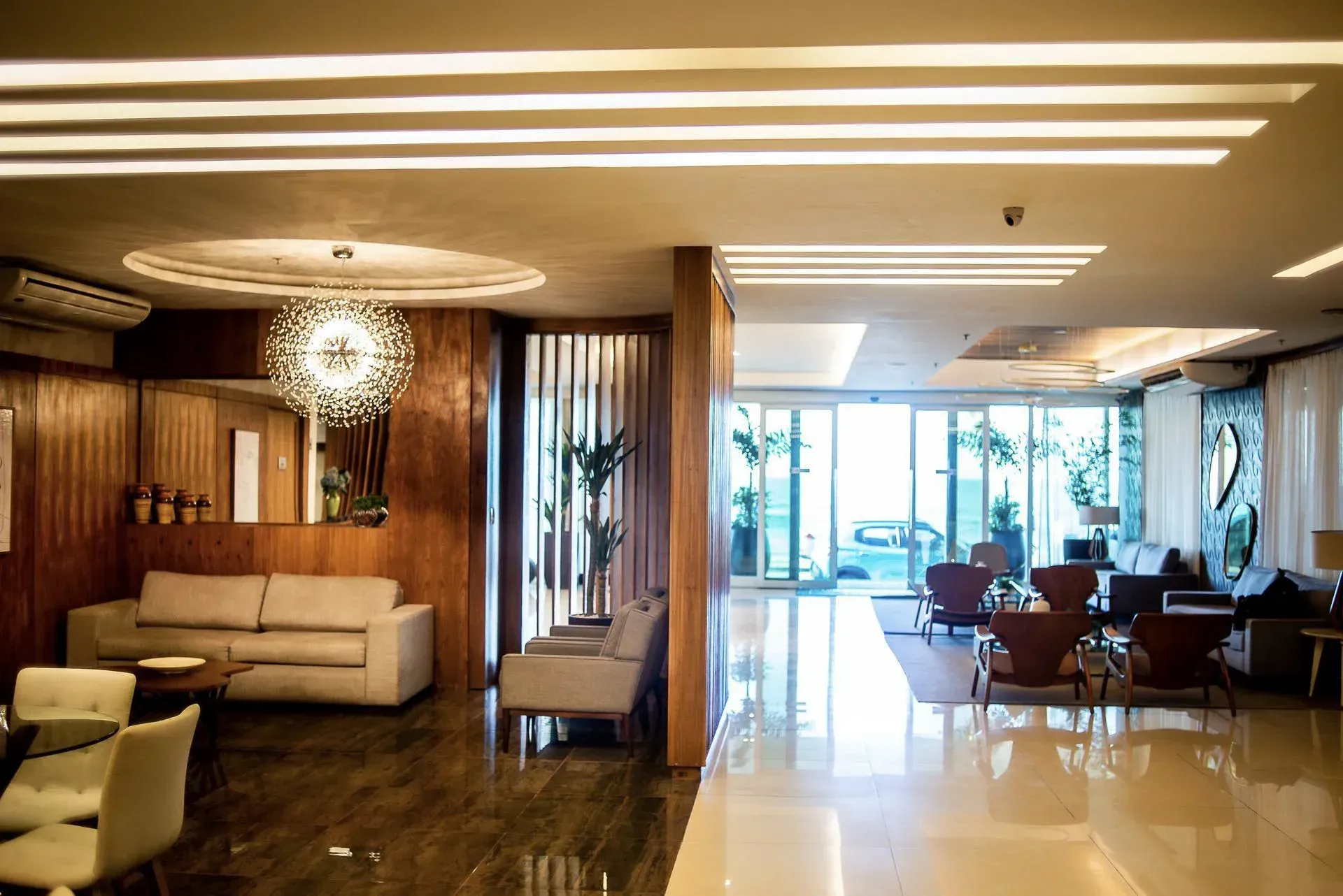 Lobby de hotel moderno com assentos, detalhes em madeira e uma área de jantar iluminada junto a grandes janelas.