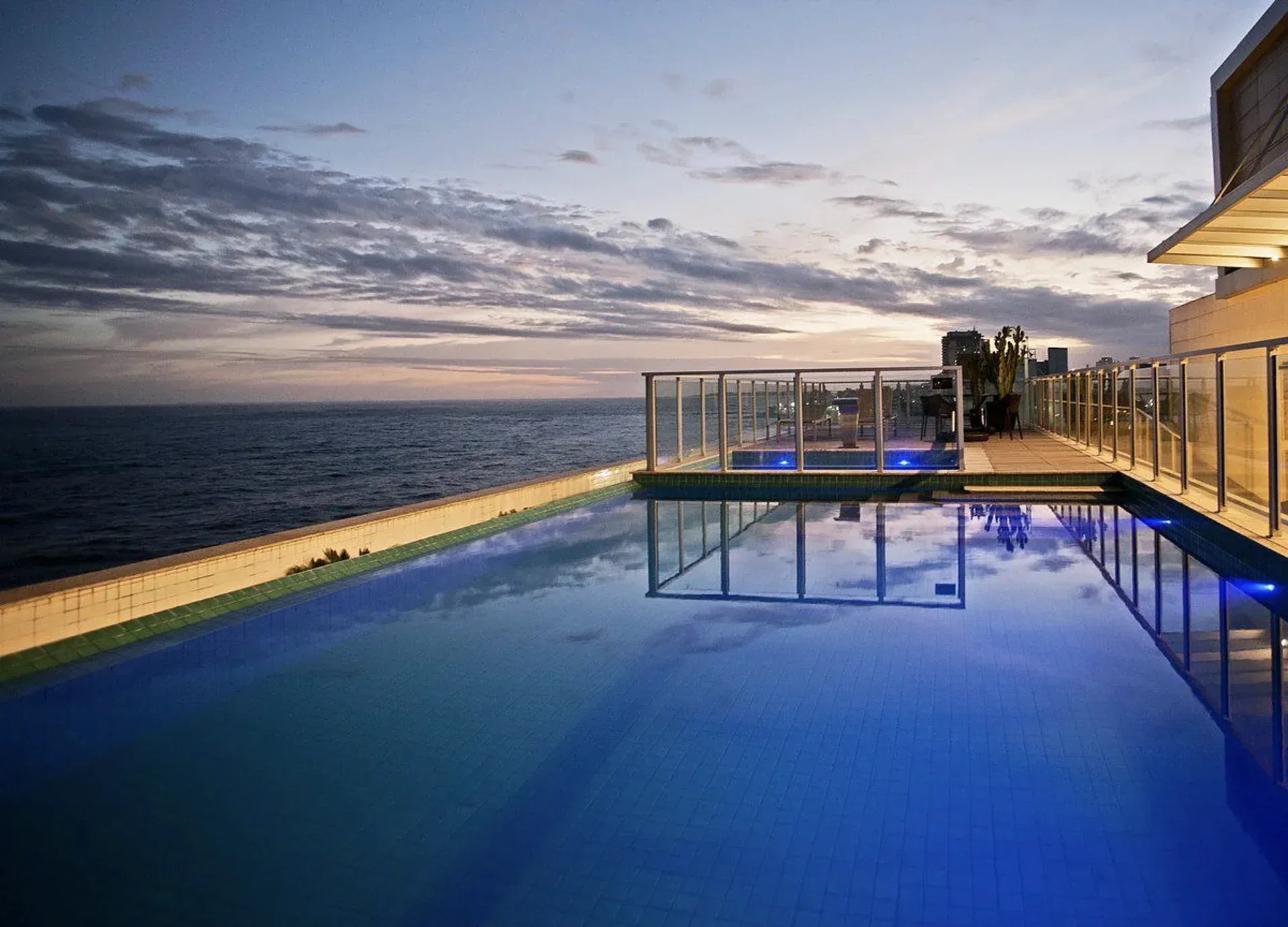 Piscina infinita à beira-mar ao entardecer, refletindo luzes azuis sob um céu nublado.