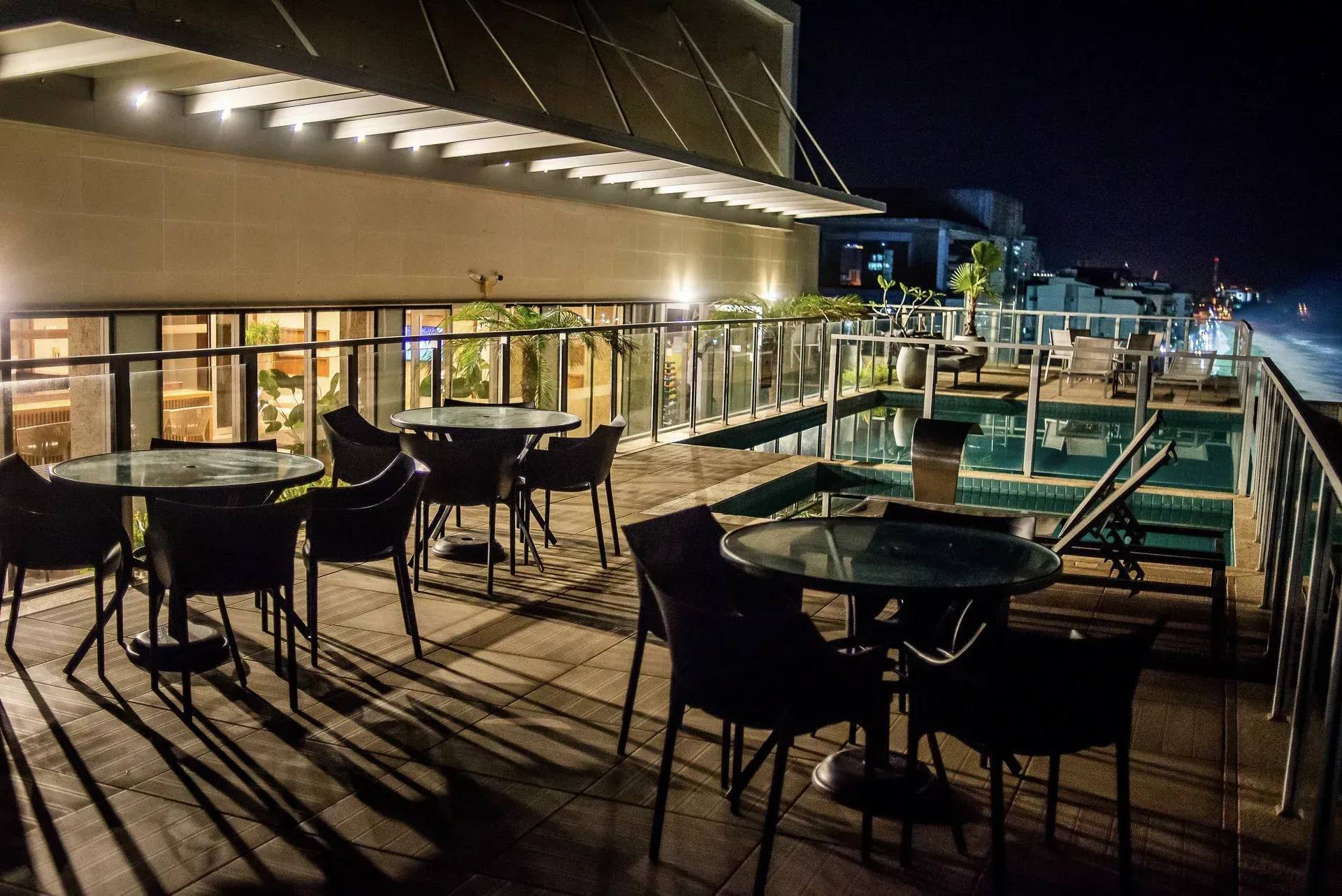 Terraço vazio de um restaurante na cobertura à noite, com mesas, cadeiras e luzes da cidade ao fundo.
