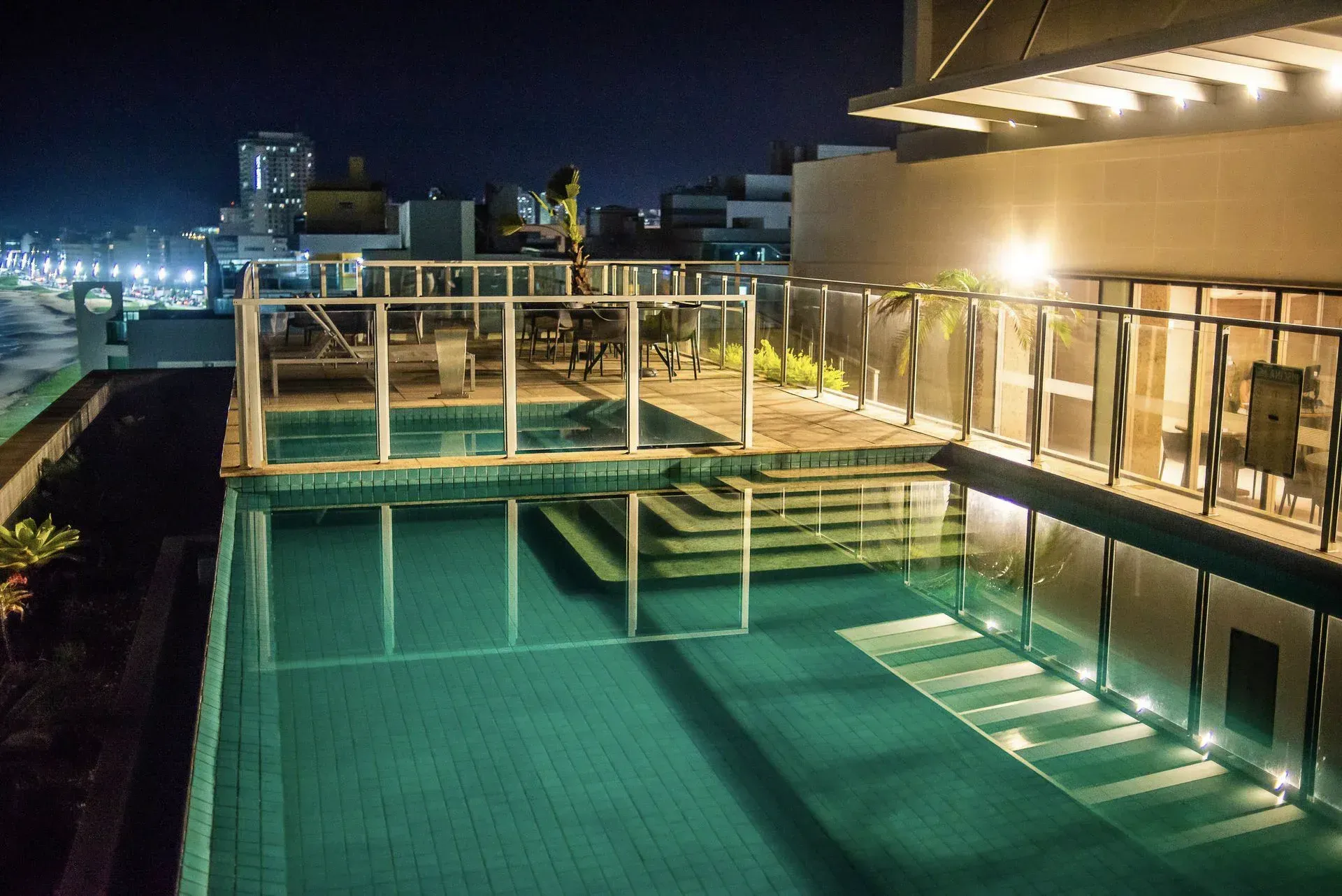 Piscina na cobertura à noite com água turquesa, guarda-corpo de vidro e luzes da cidade ao fundo.