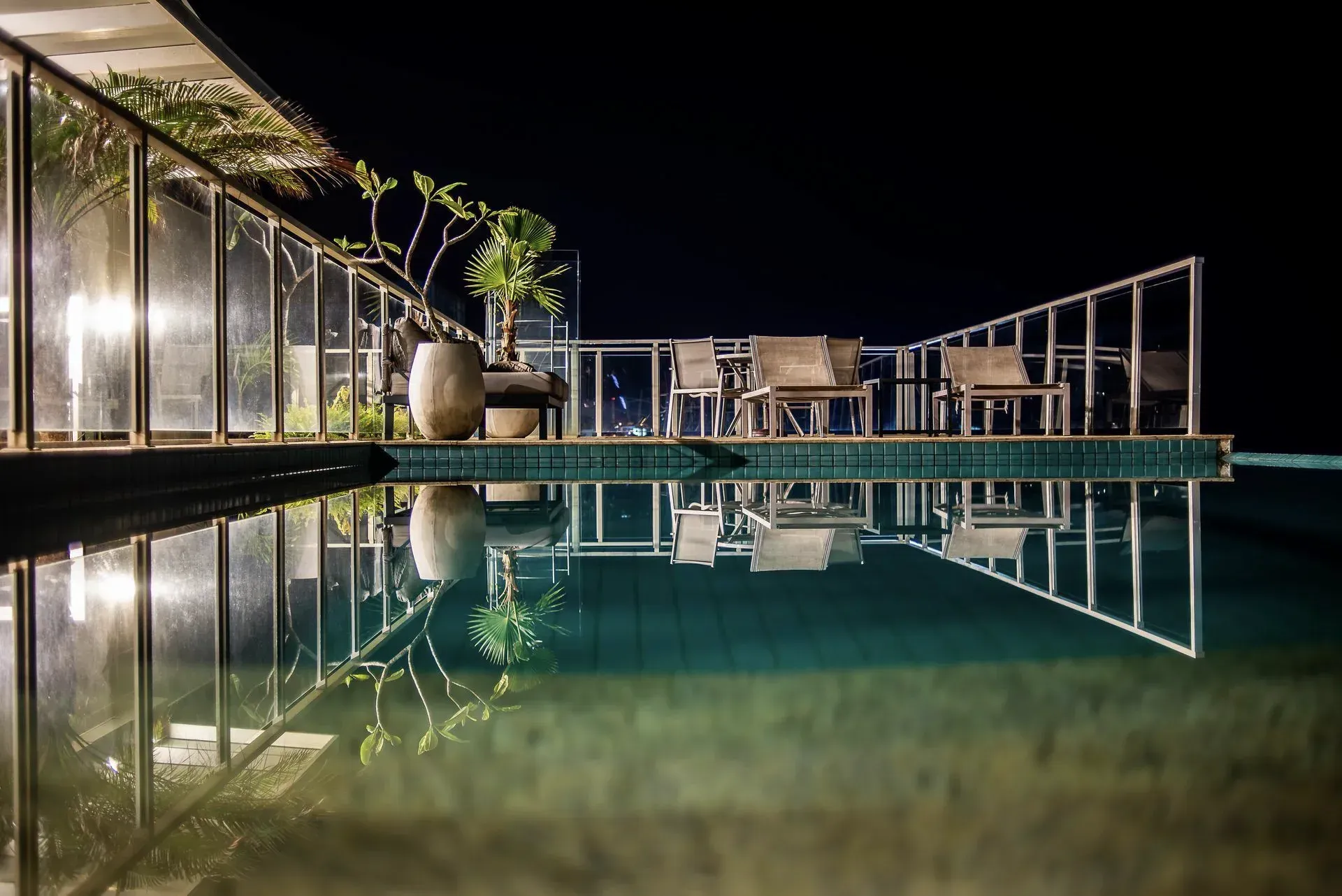 Deck noturno à beira da piscina com guarda-corpos de vidro, espreguiçadeiras e o reflexo da água tranquila.