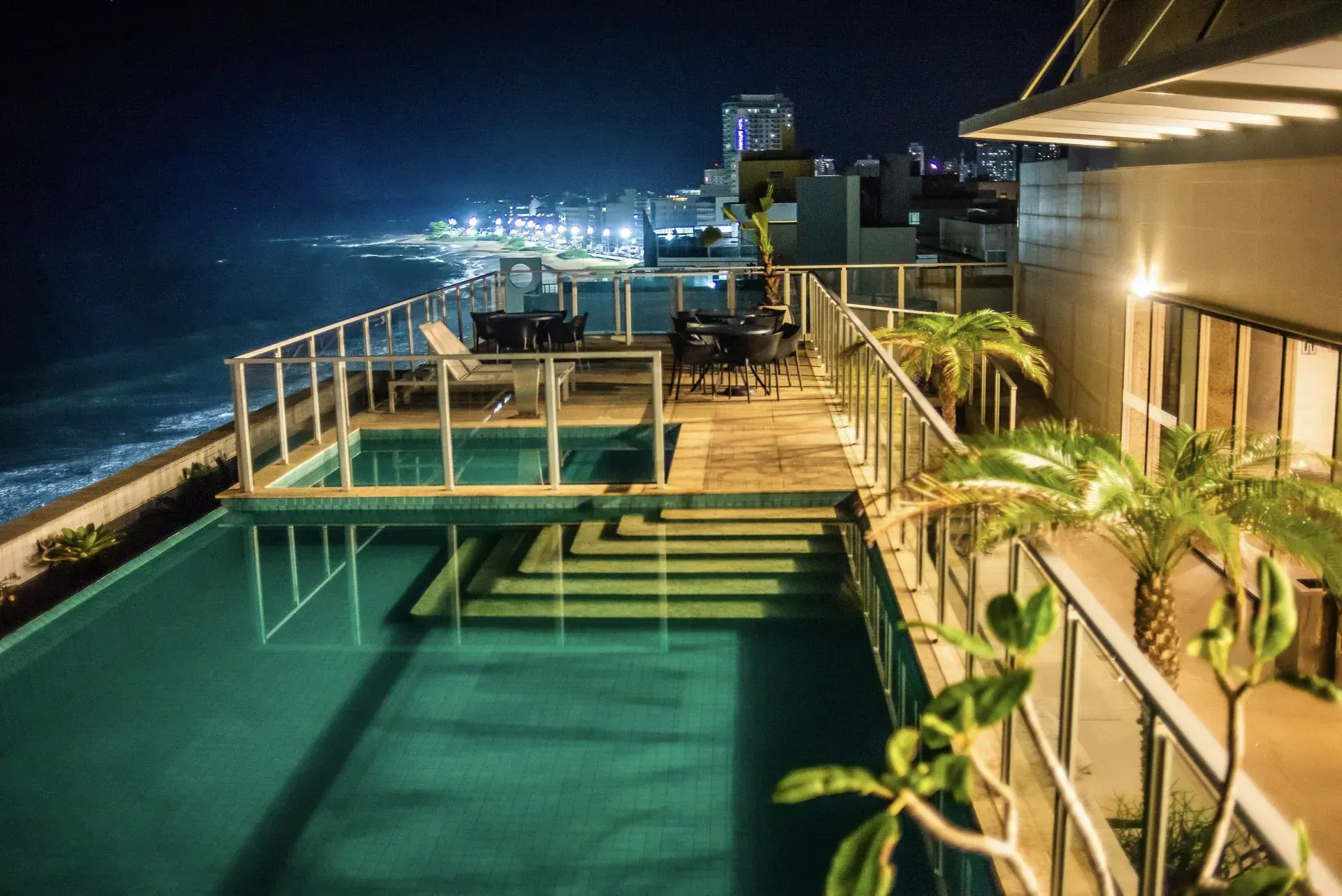 Deck da piscina à beira-mar à noite, com espreguiçadeiras e luzes da cidade ao longo da costa.