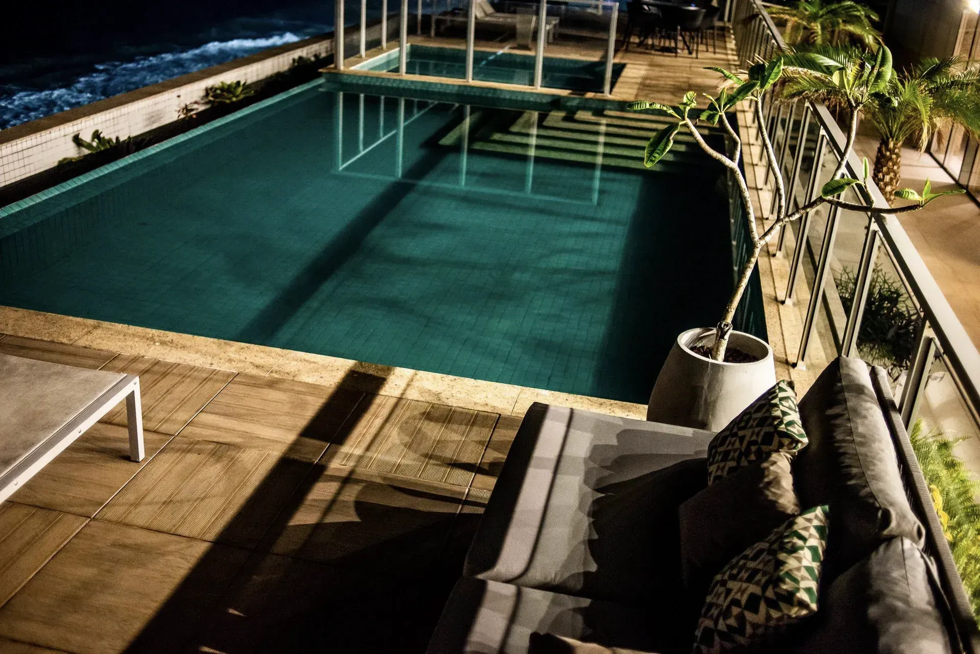 Vista noturna de uma piscina externa iluminada ao lado de um deck de madeira e área de estar à beira-mar.