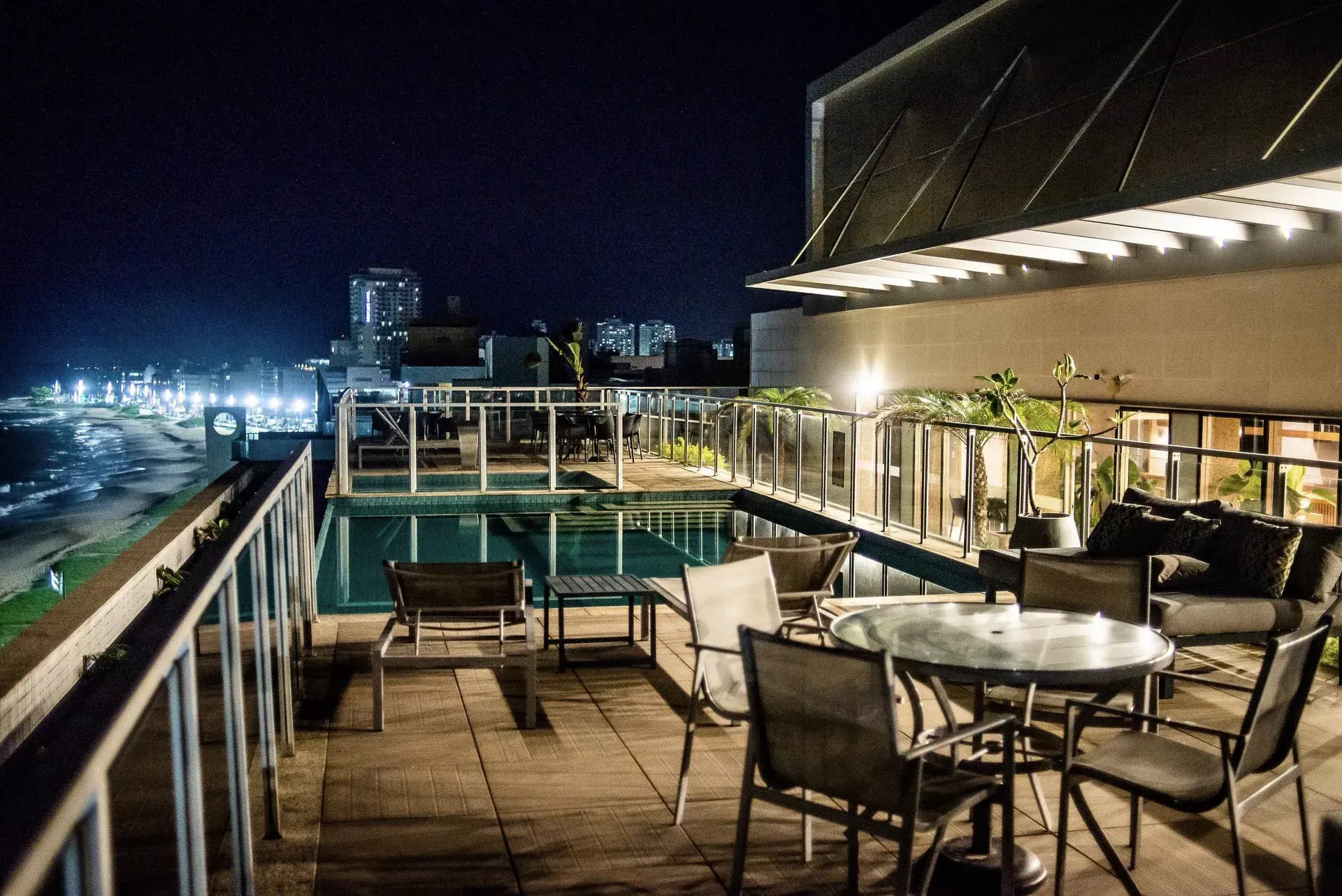 Terraço da piscina na cobertura à noite, com espreguiçadeiras, mesas e luzes da cidade além do parapeito.