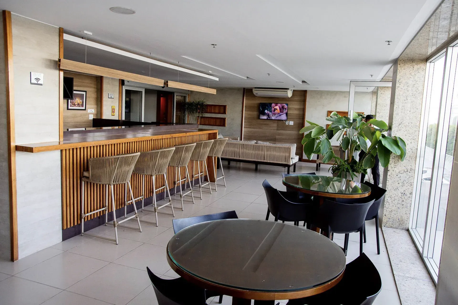 Salão de estar moderno com banquetas de bar, mesas redondas, plantas e janelas amplas; TV na parede do fundo.