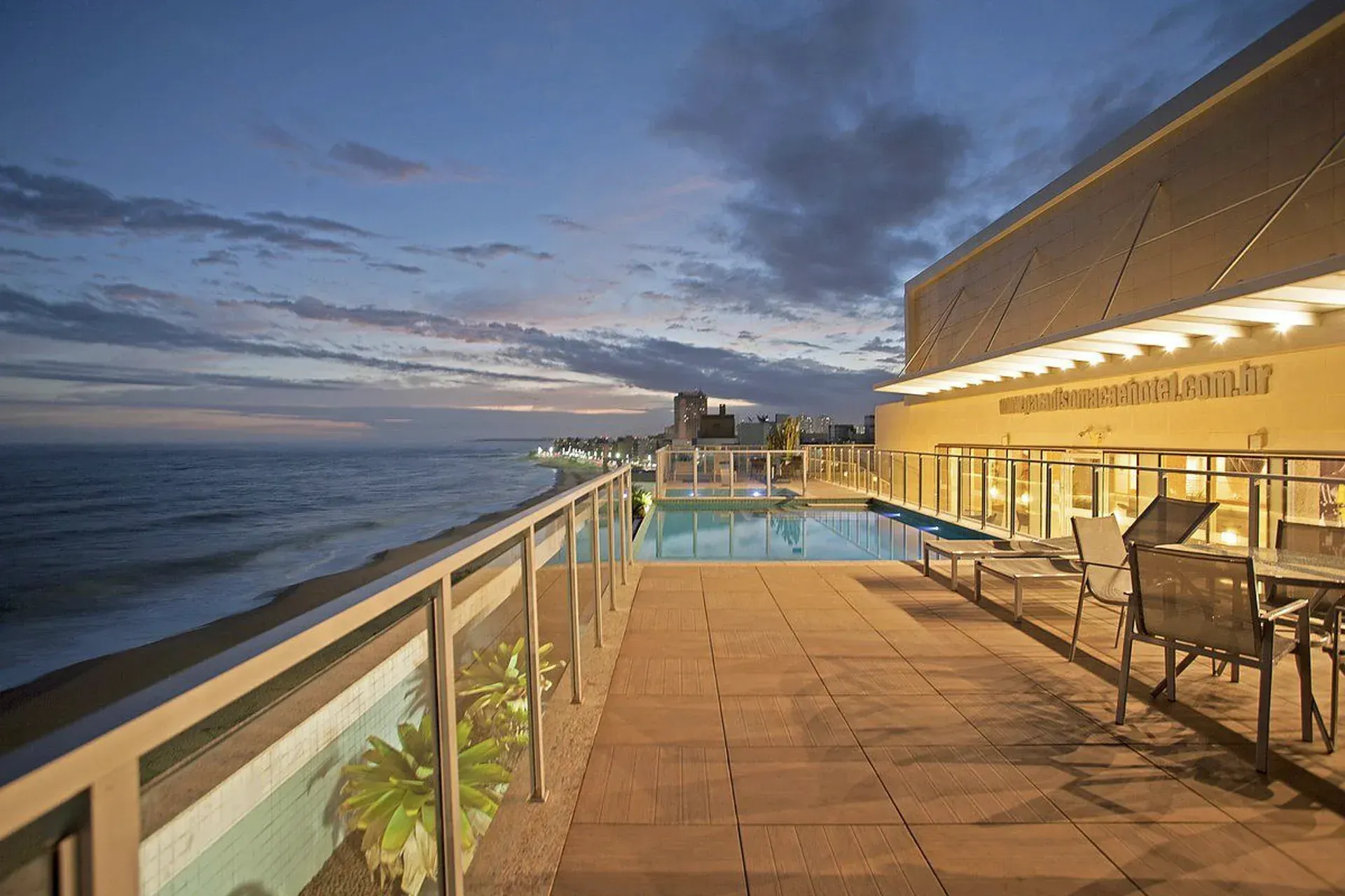 Terraço à beira-mar com piscina iluminada ao entardecer, com vista para o mar sob um céu nublado.