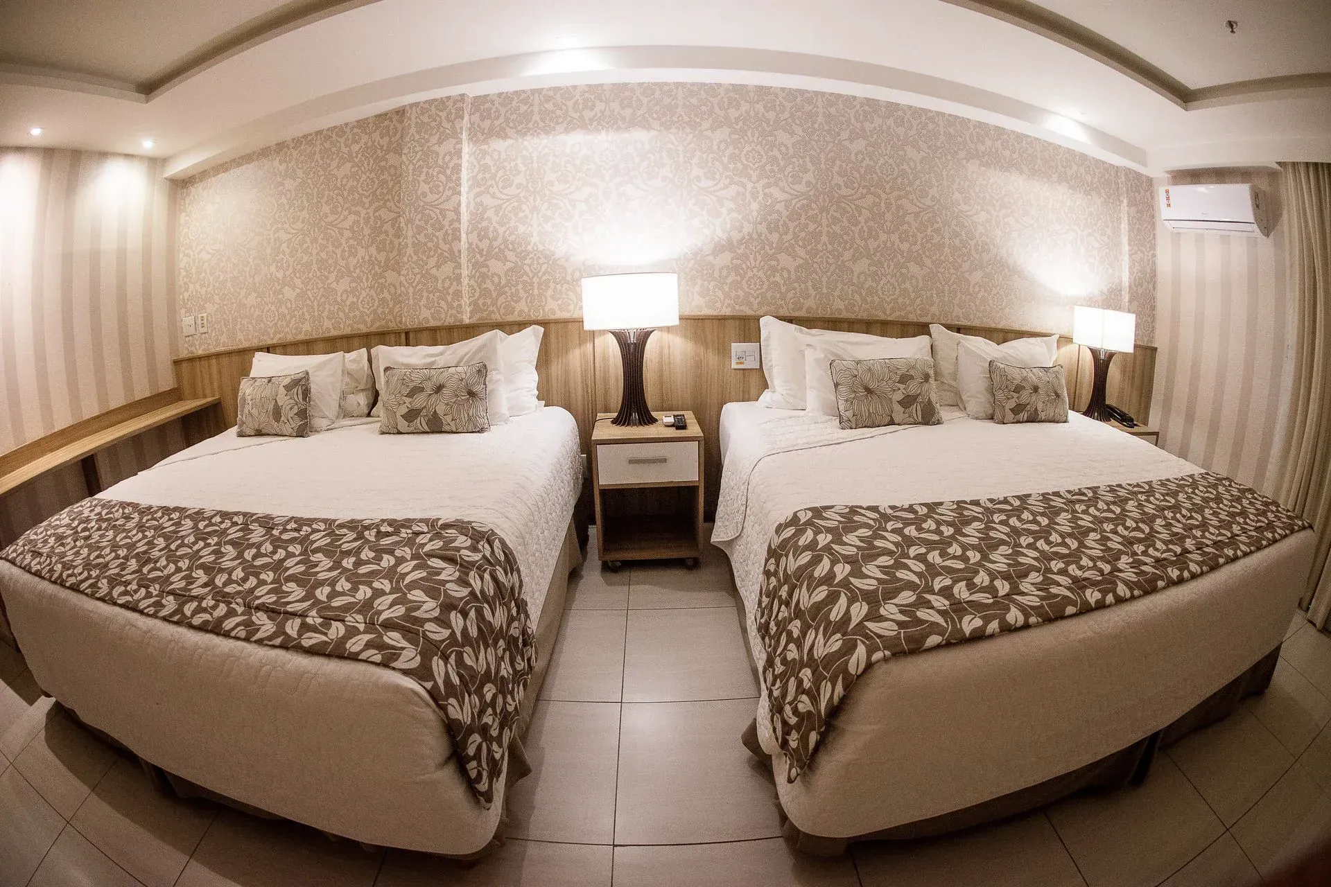Quarto de hotel com duas camas de solteiro, peseiras estampadas e um abajur entre elas.