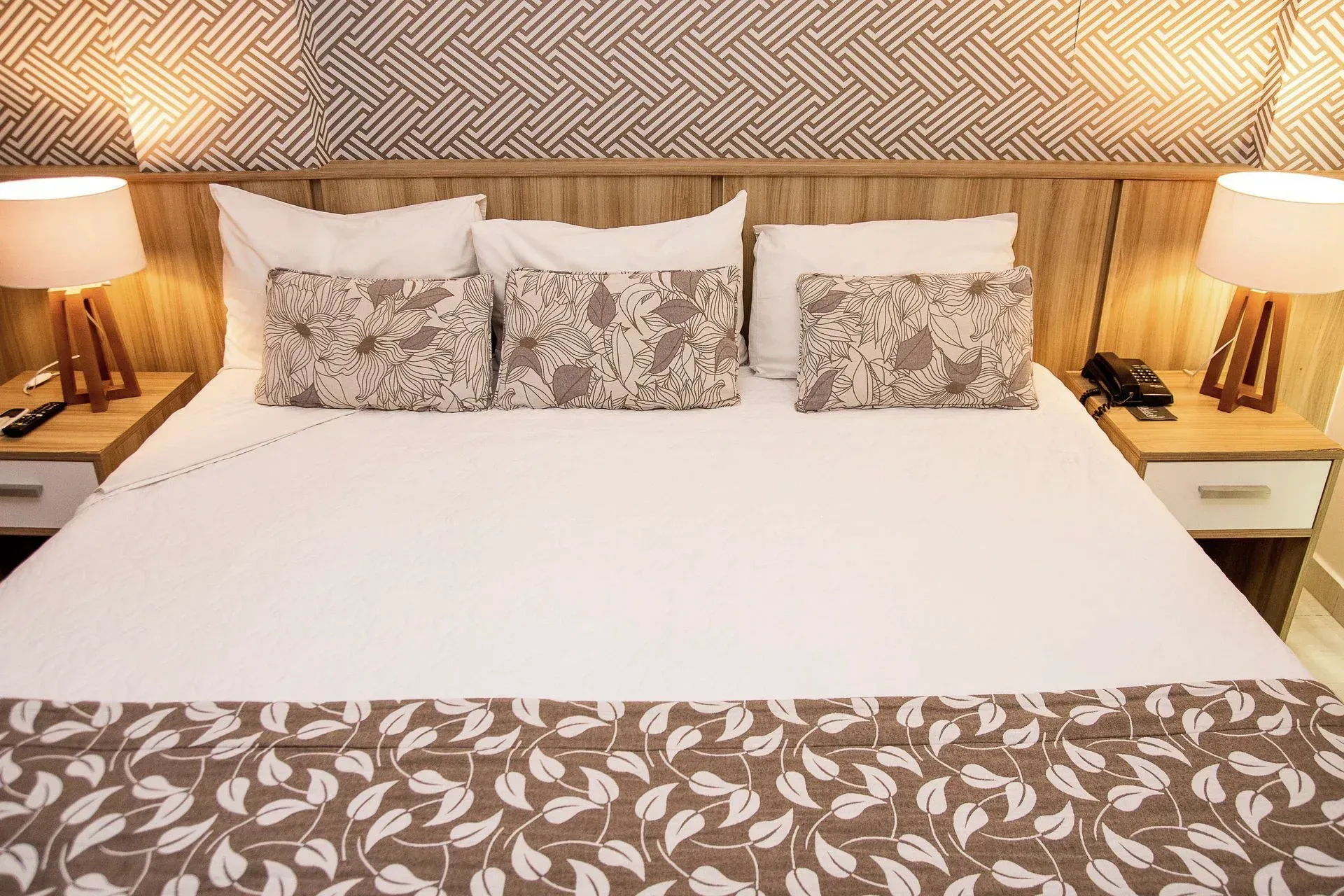 Cama arrumada com lençóis brancos, almofadas estampadas, abajures e cabeceira de vime em um quarto aconchegante.