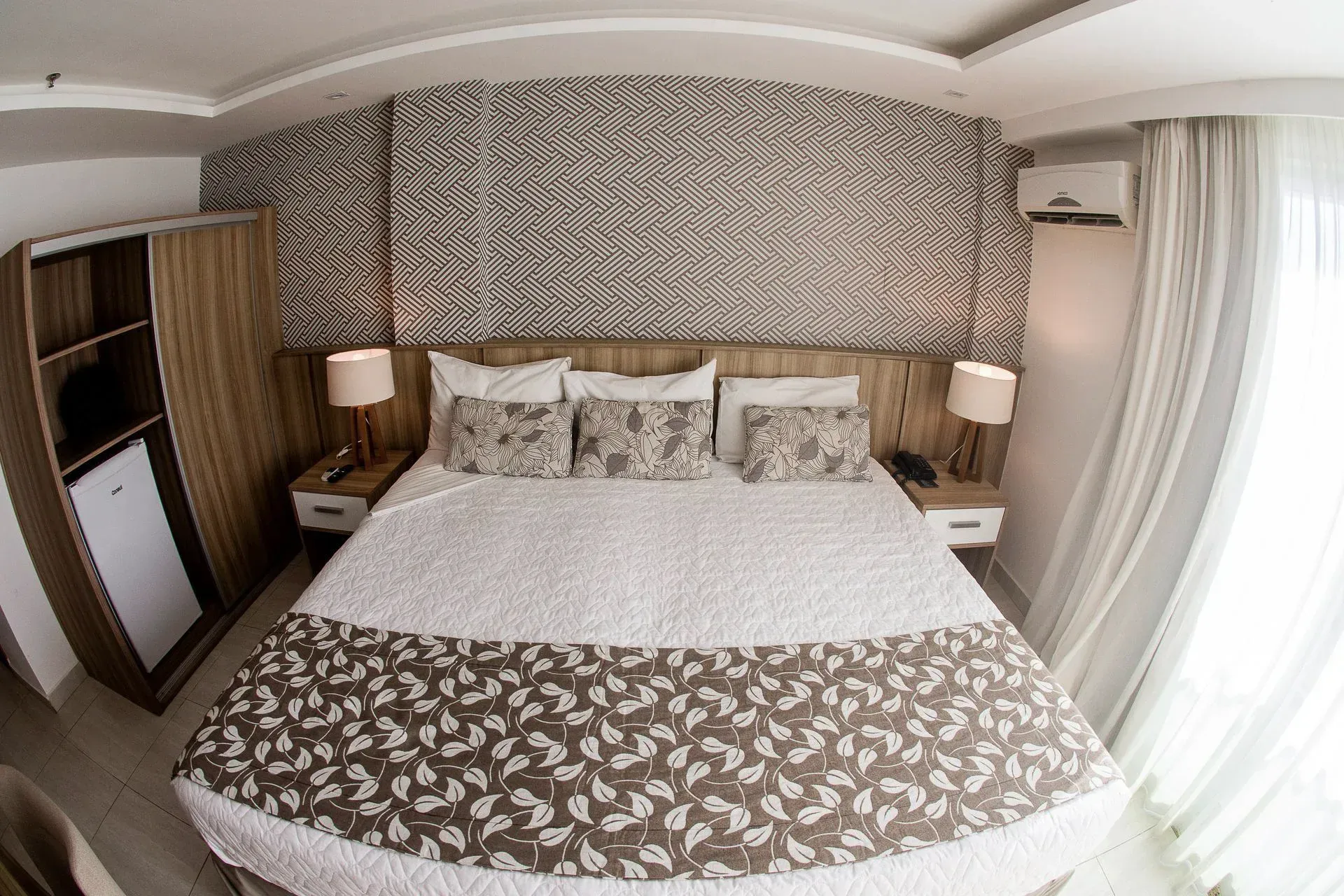 Cama de hotel impecavelmente arrumada com papel de parede estampado, abajures e cortinas do chão ao teto.