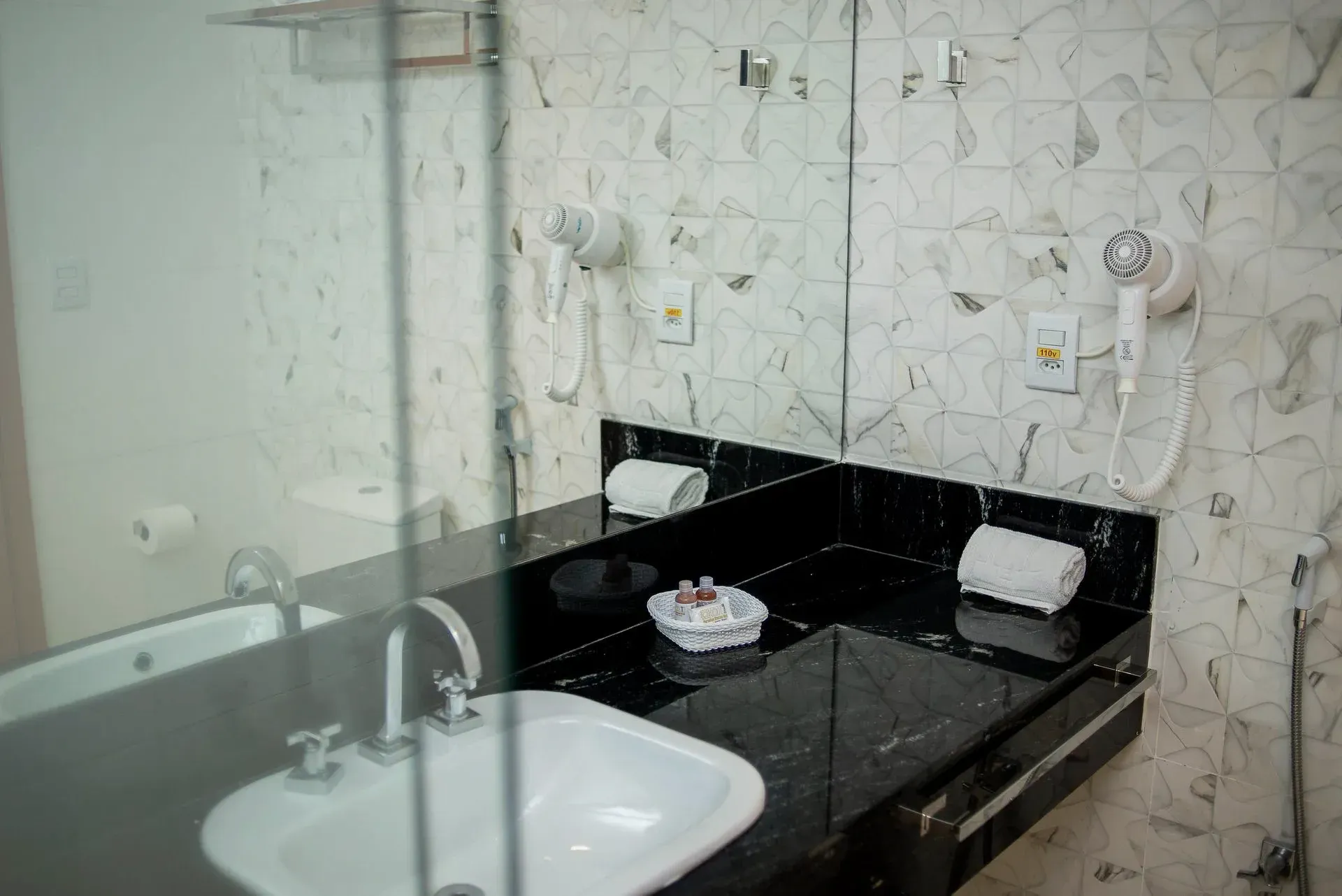 Banheiro moderno com box de vidro, pia e bancada de mármore preto com artigos de higiene pessoal.