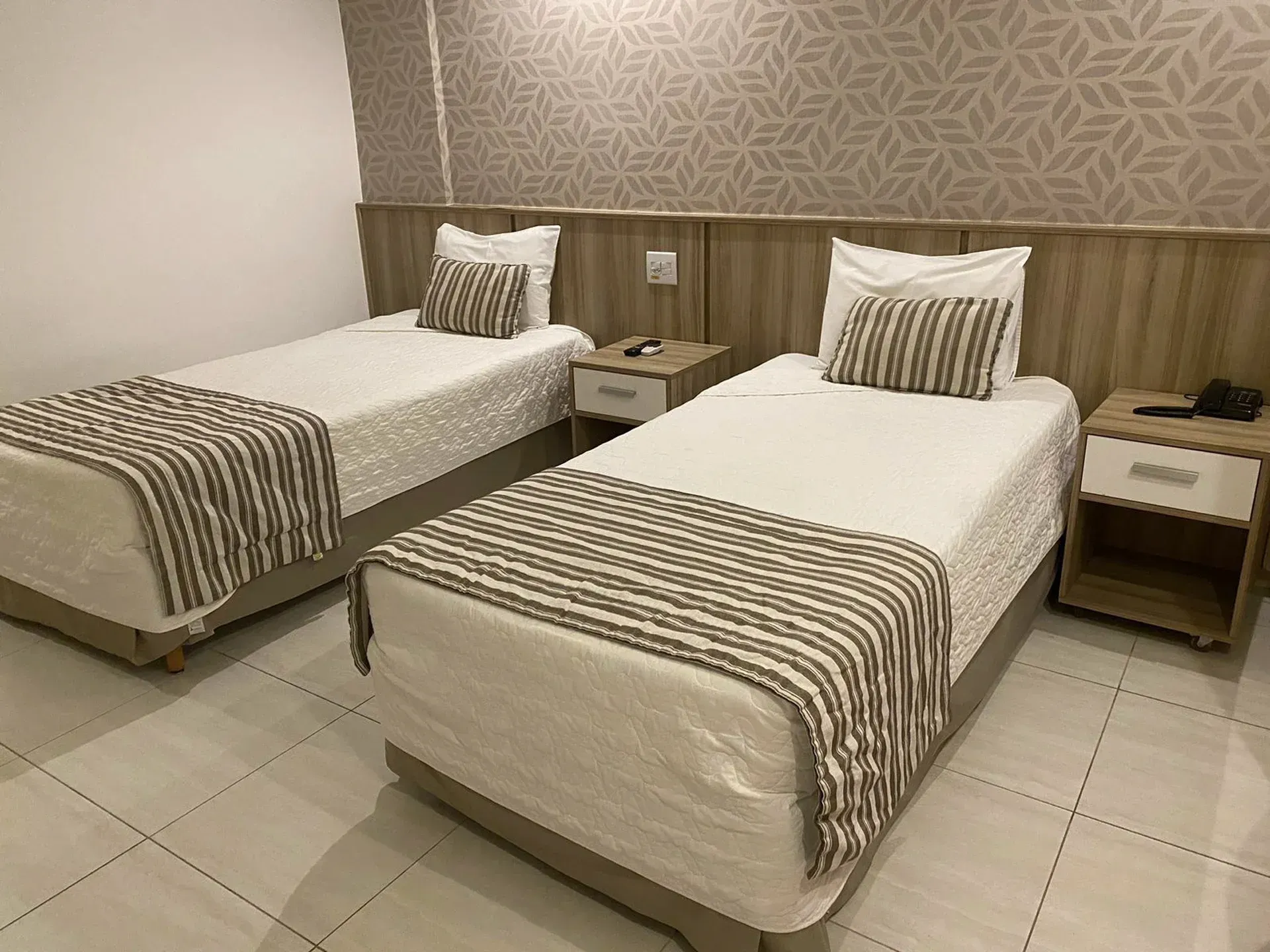 Quarto de hotel com duas camas de solteiro, roupa de cama listrada e mesas de cabeceira sobre piso de azulejo.