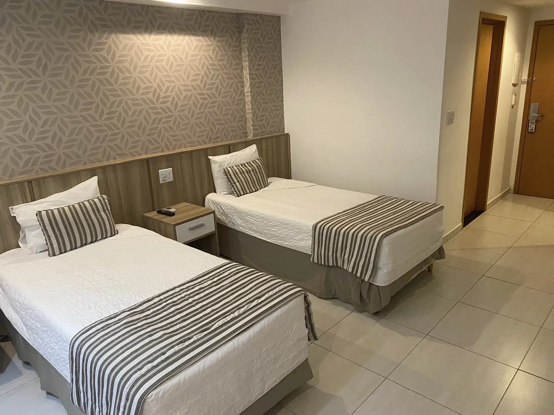 Quarto de hotel com duas camas de solteiro, peseira listrada, mesa de cabeceira e piso de azulejo.
