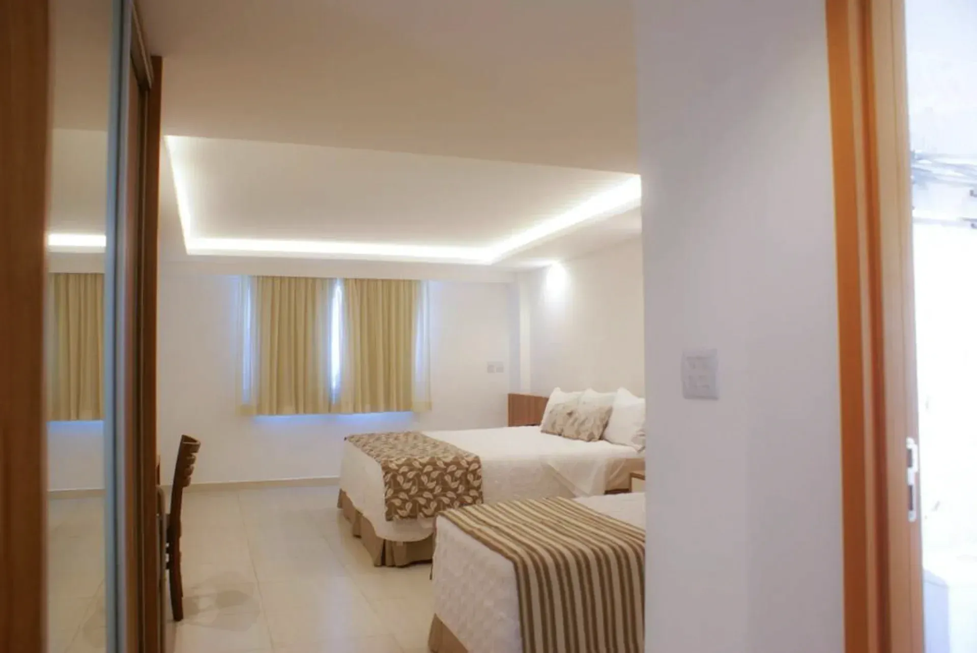 Quarto moderno e luminoso com paredes brancas, roupa de cama bege, pufe listrado e iluminação suave no teto.