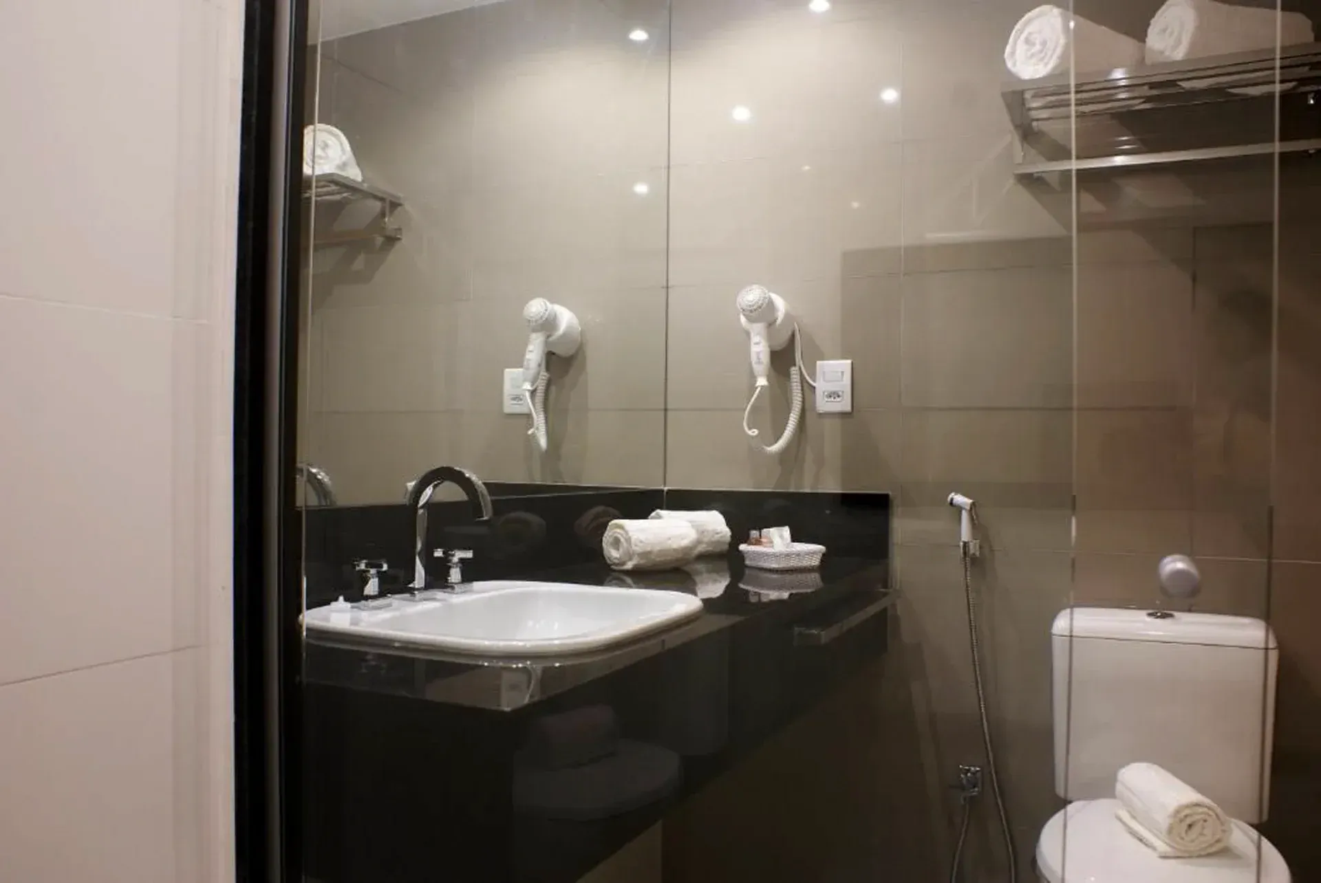 Banheiro moderno com pia, espelho, secadores de cabelo fixados na parede e vaso sanitário atrás de uma divisória de vidro.