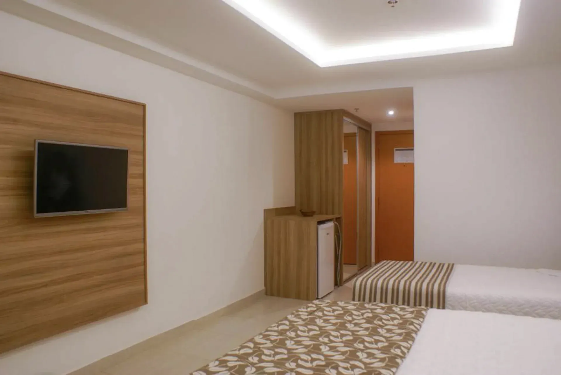 Quarto de hotel moderno com TV de parede, duas camas de solteiro, detalhes em madeira e iluminação aconchegante.