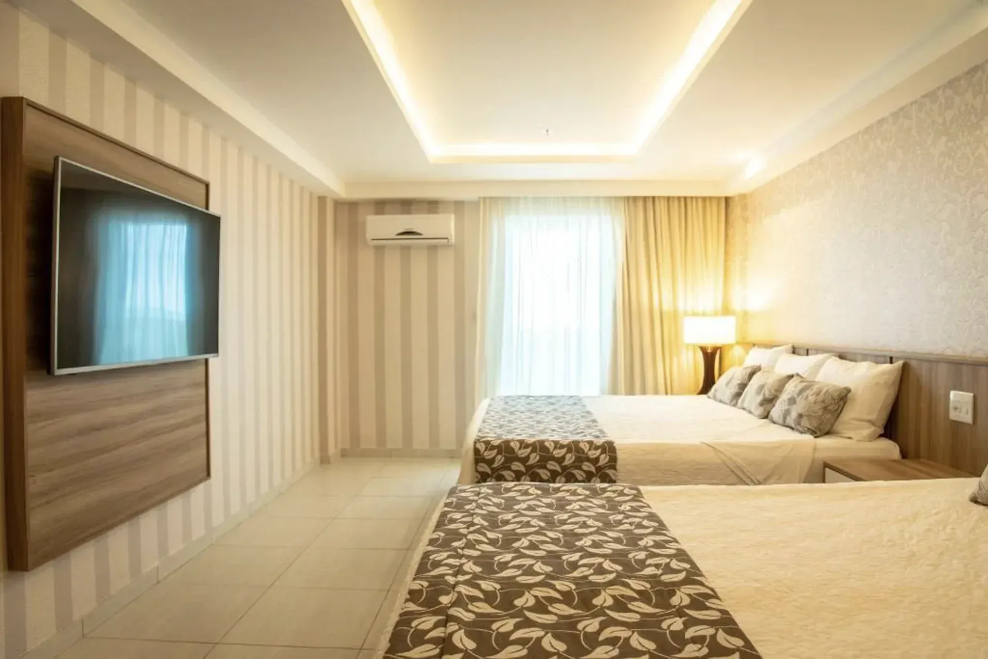 Quarto de hotel moderno em tons de bege com duas camas, TV de parede, papel de parede listrado e uma janela bem iluminada.