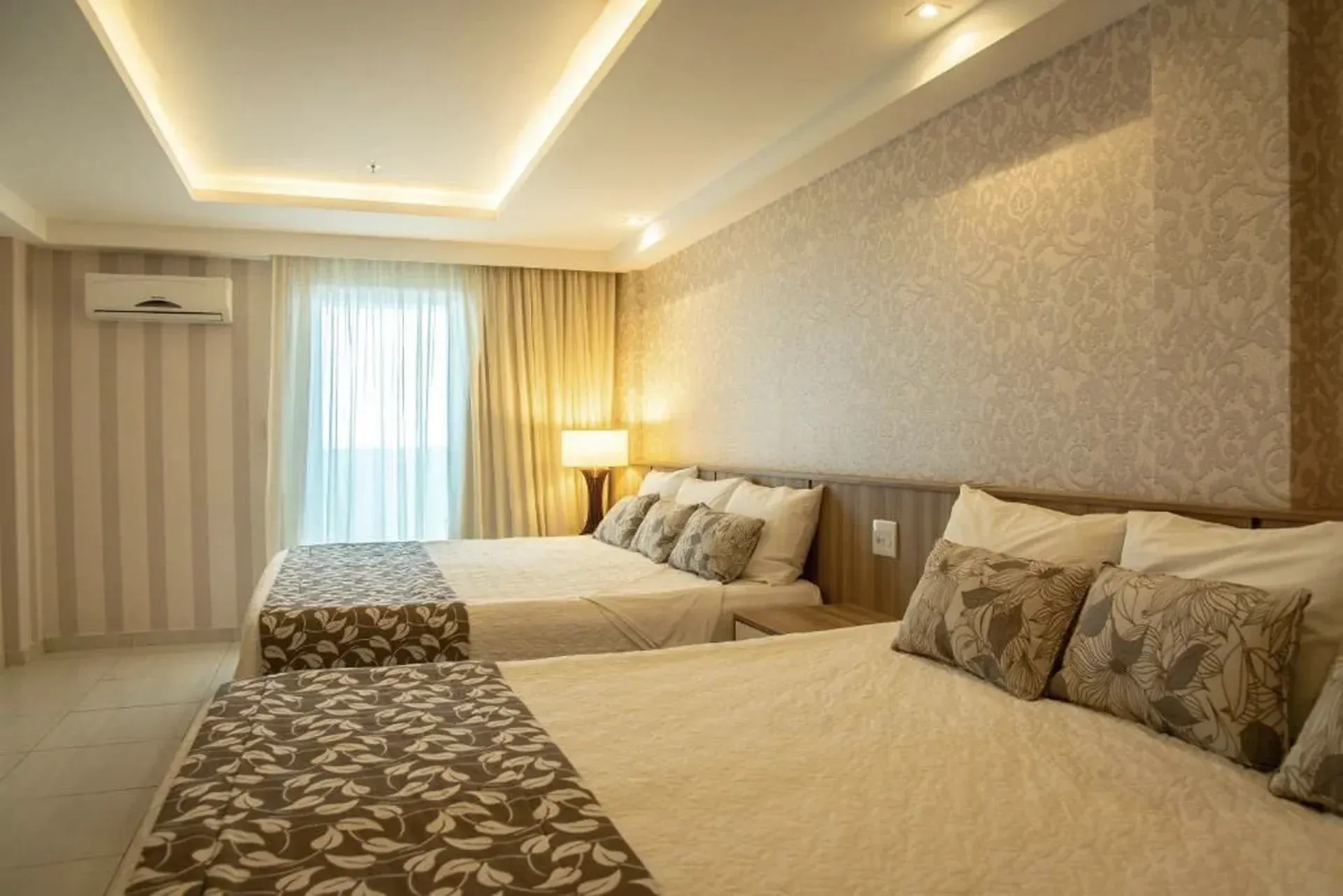 Quarto de solteiro moderno com duas camas de solteiro, decoração em tons de bege, colchas estampadas, cortinas e iluminação.