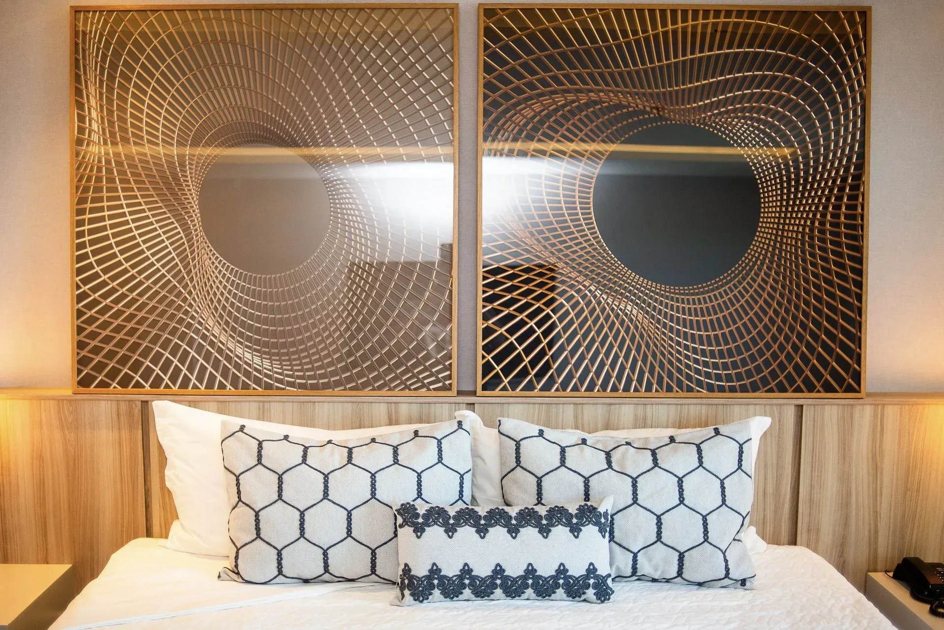 Cama de hotel com travesseiros hexagonais brancos sob uma obra de arte geométrica dourada na parede e painéis de madeira.