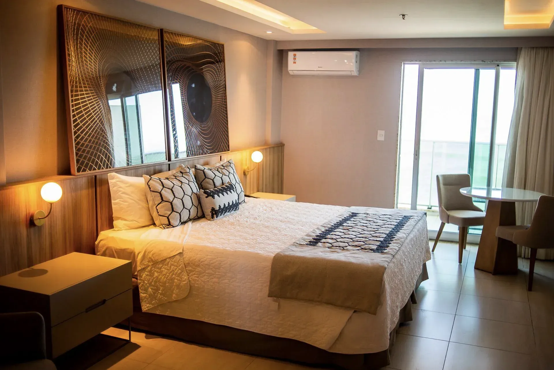 Quarto de hotel moderno com cama king-size, abajures, portas para varanda e uma pequena mesa com cadeiras junto à janela.