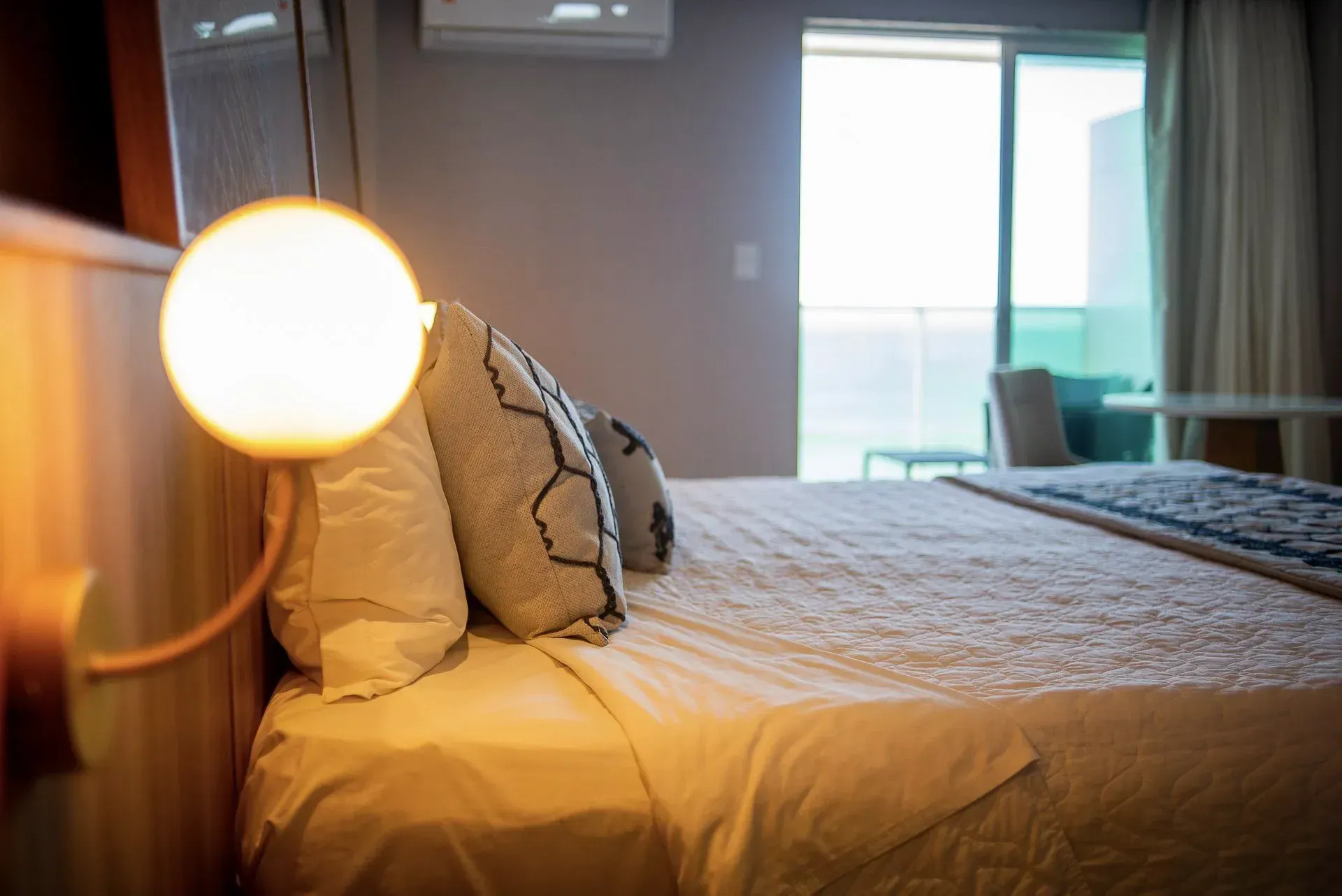 Quarto aconchegante com cama arrumada, abajur redondo em primeiro plano e janela iluminada junto à varanda.